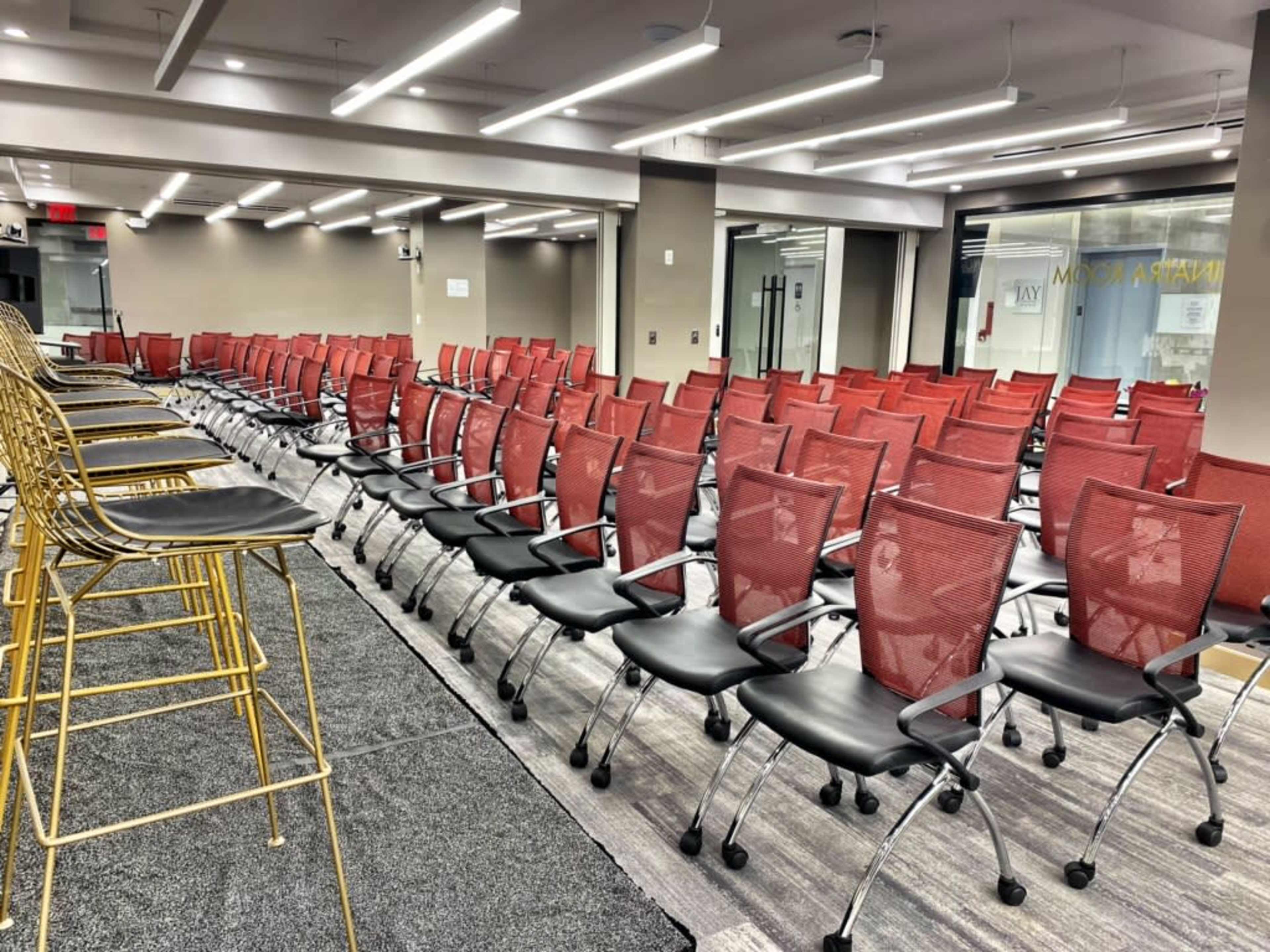 A conference room is set up with rows of red mesh chairs and several gold wire stools positioned at the front.