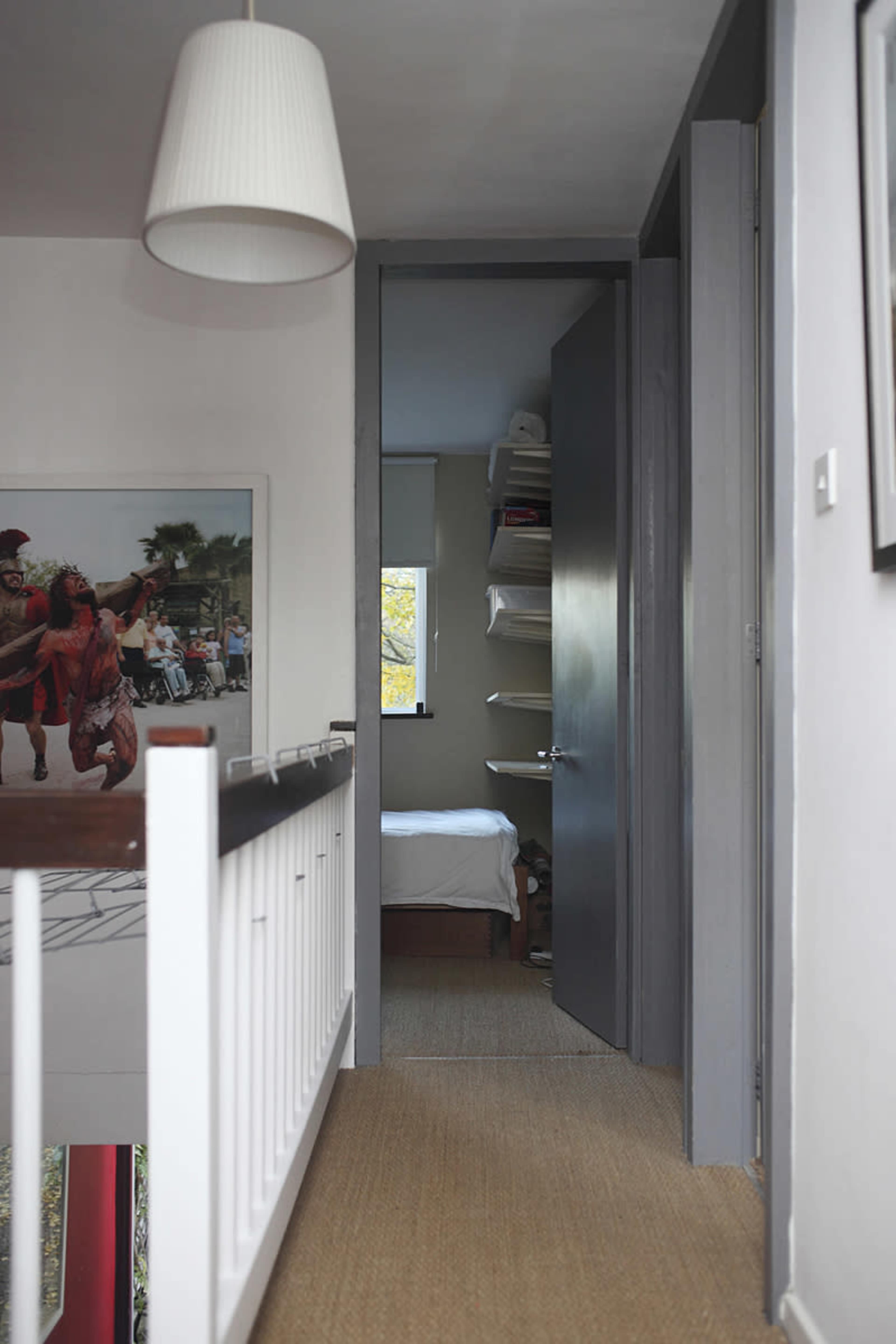 A hallway with a pendant light leads to a doorway opening into a small bedroom, with a shelf and a window visible.