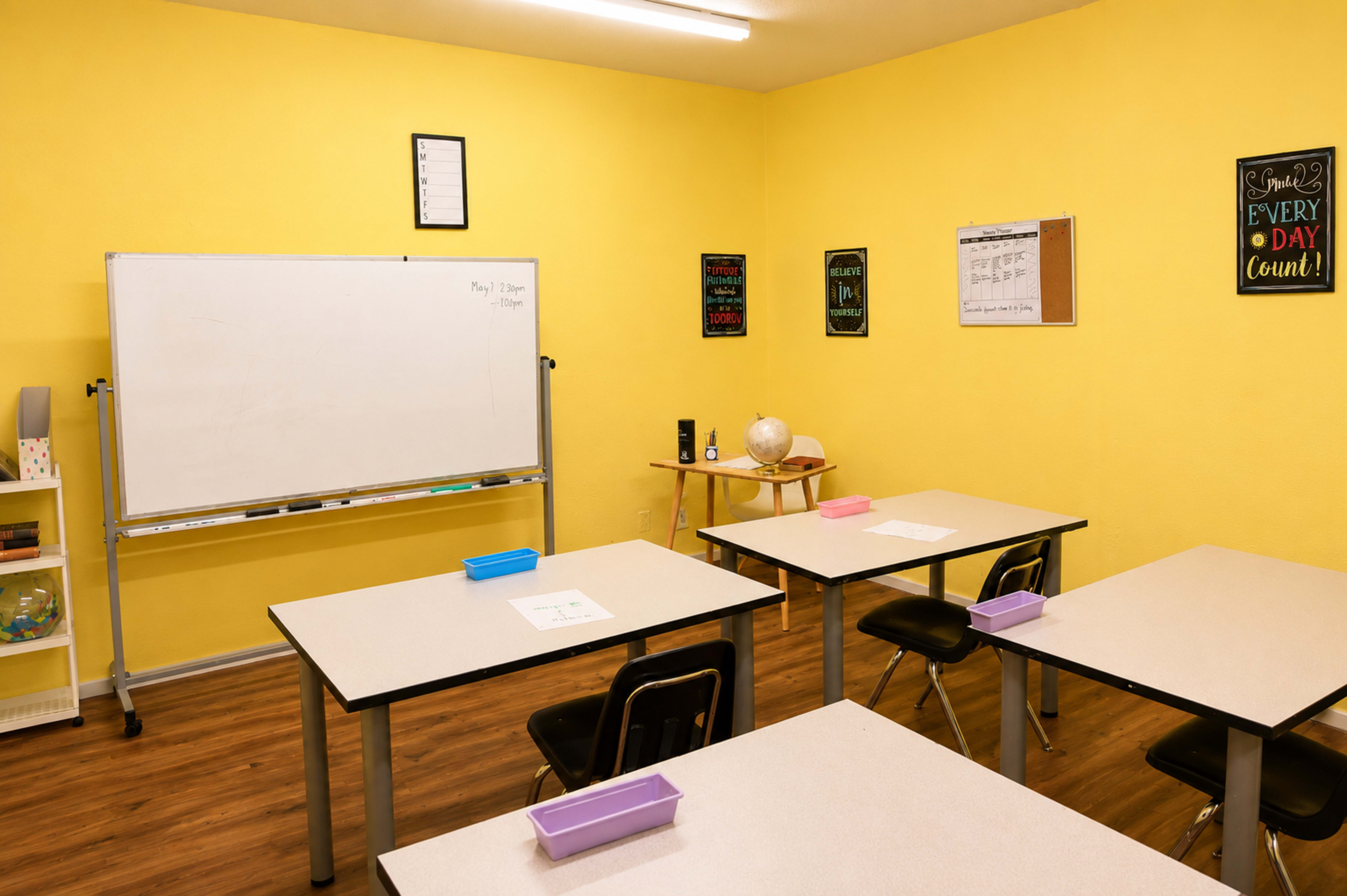 A brightly colored classroom features a whiteboard, wooden tables with black chairs, and educational posters on the walls.