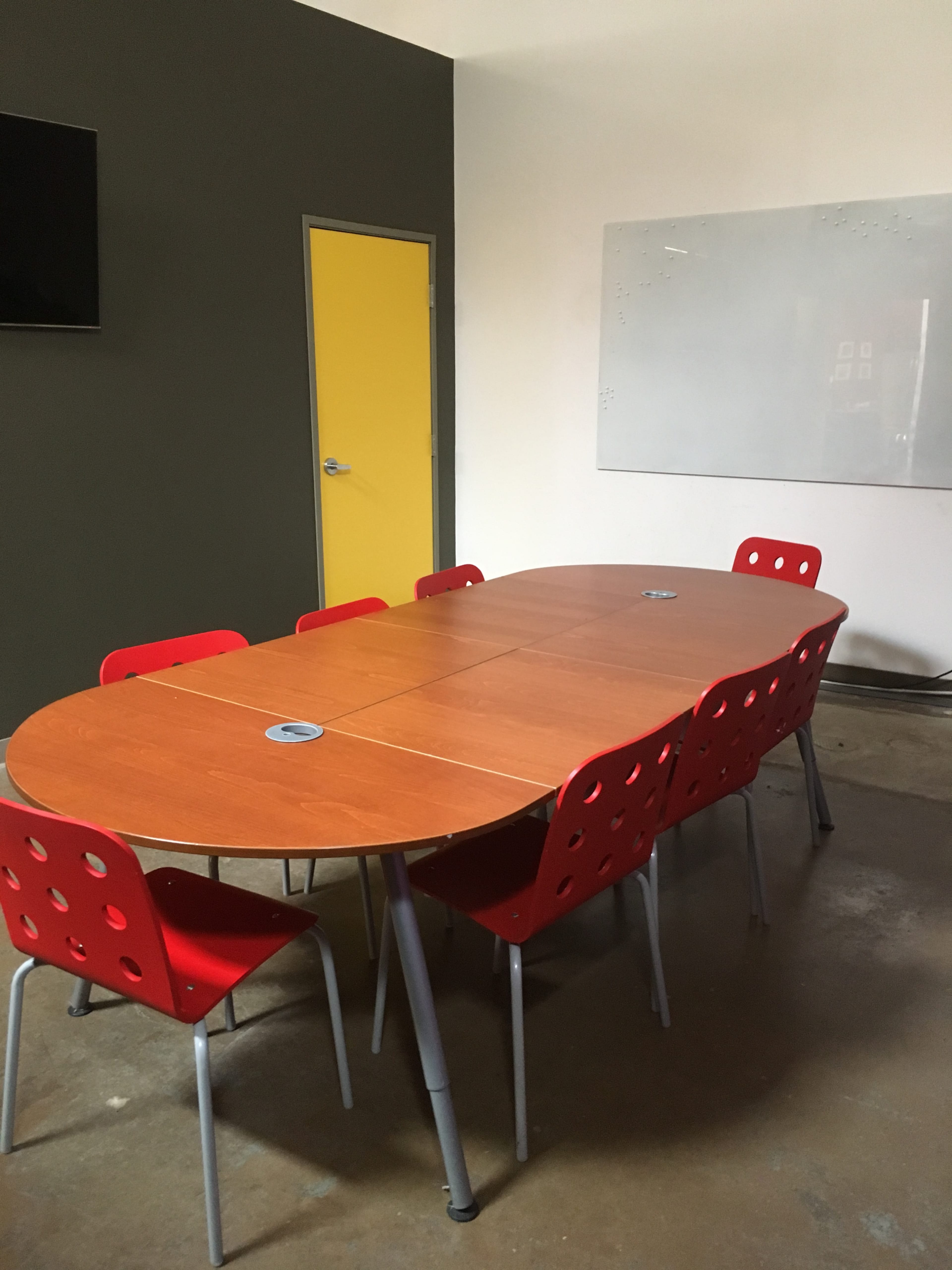 The image shows a conference room with a large oval table surrounded by red chairs, a whiteboard on one wall, and a yellow door.