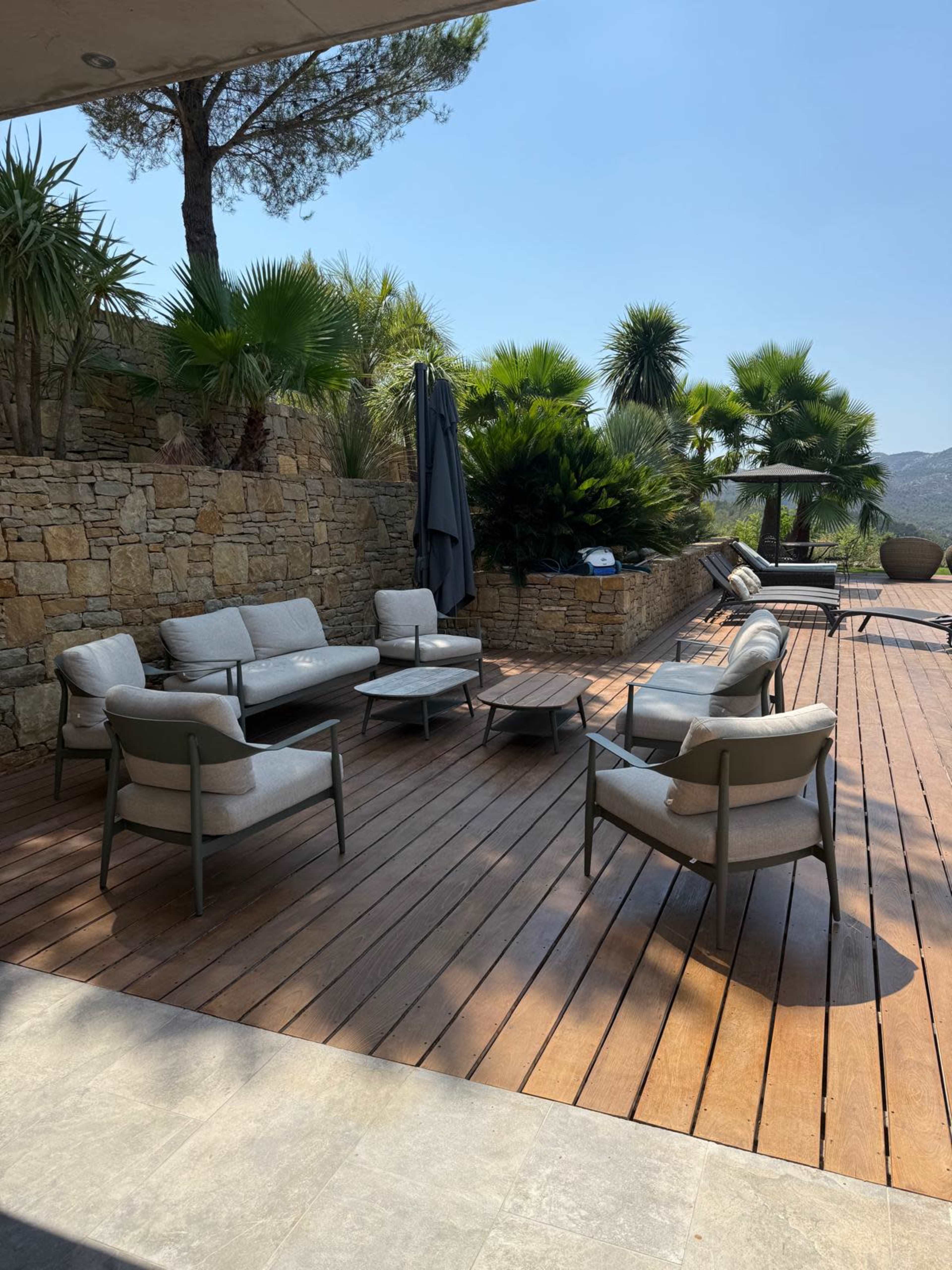 The image shows a patio with modern outdoor furniture arranged on wooden decking, surrounded by lush greenery and palm trees.