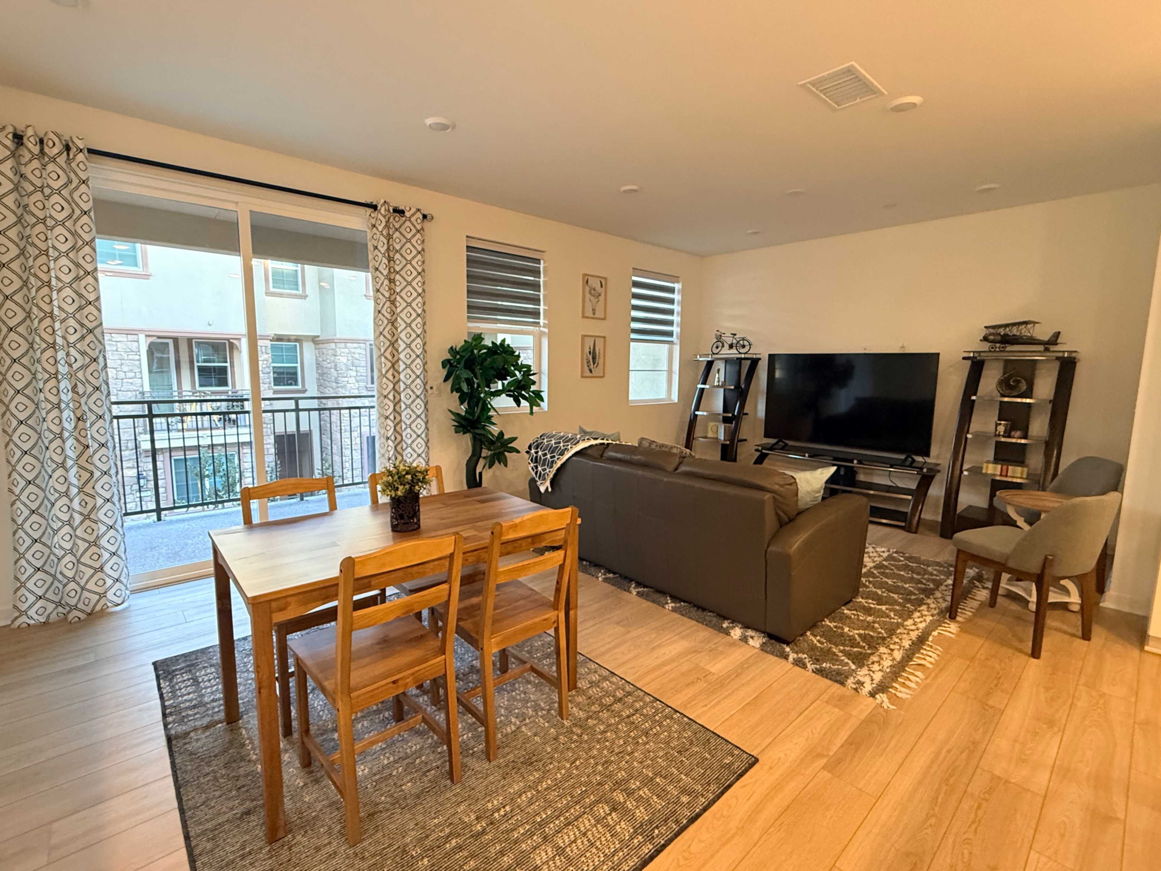 The image shows a modern living area with a dining table, chairs, a couch, a television, and large windows allowing natural light.