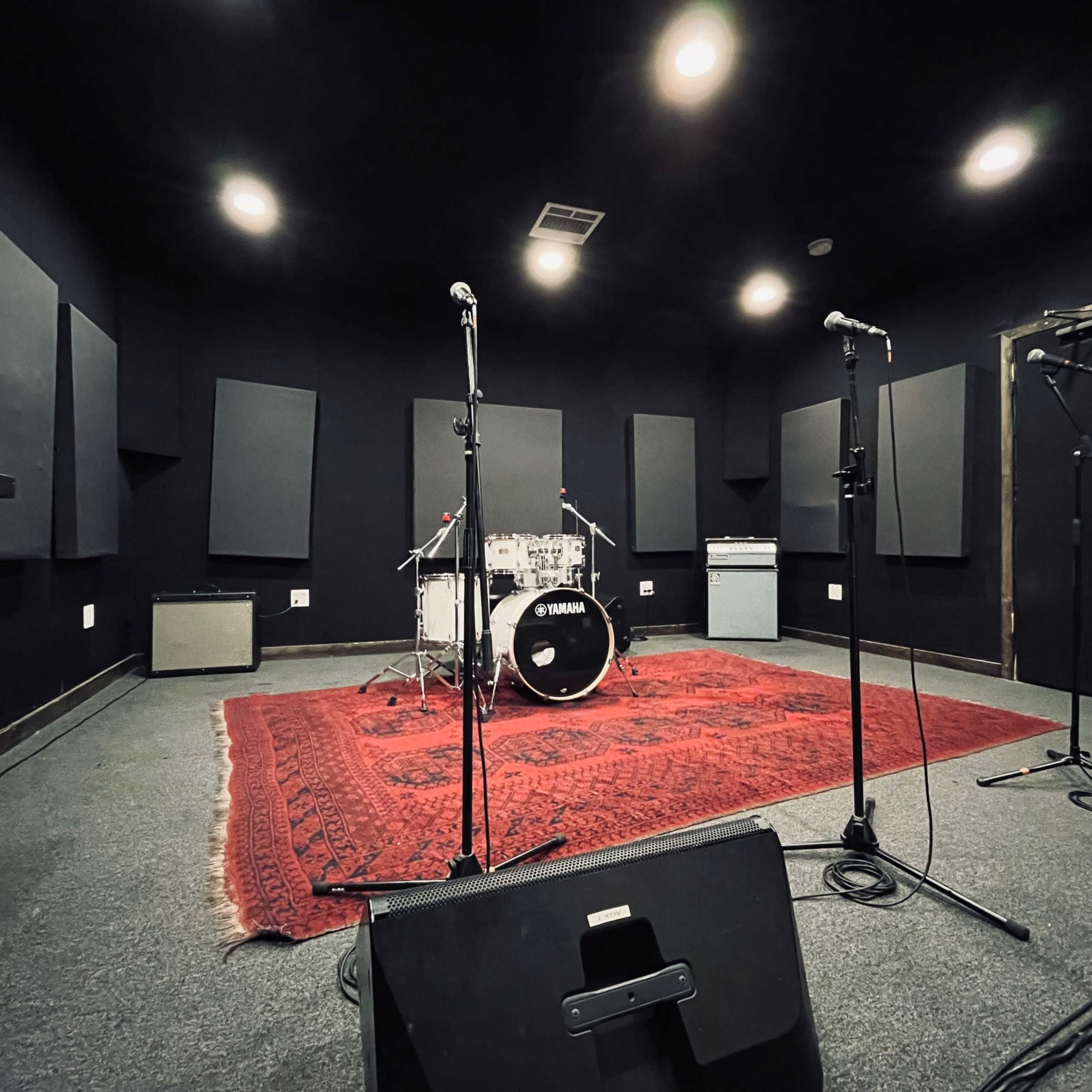 The image shows a music rehearsal space with a drum set positioned on a red rug, flanked by microphones and soundproof wall panels.