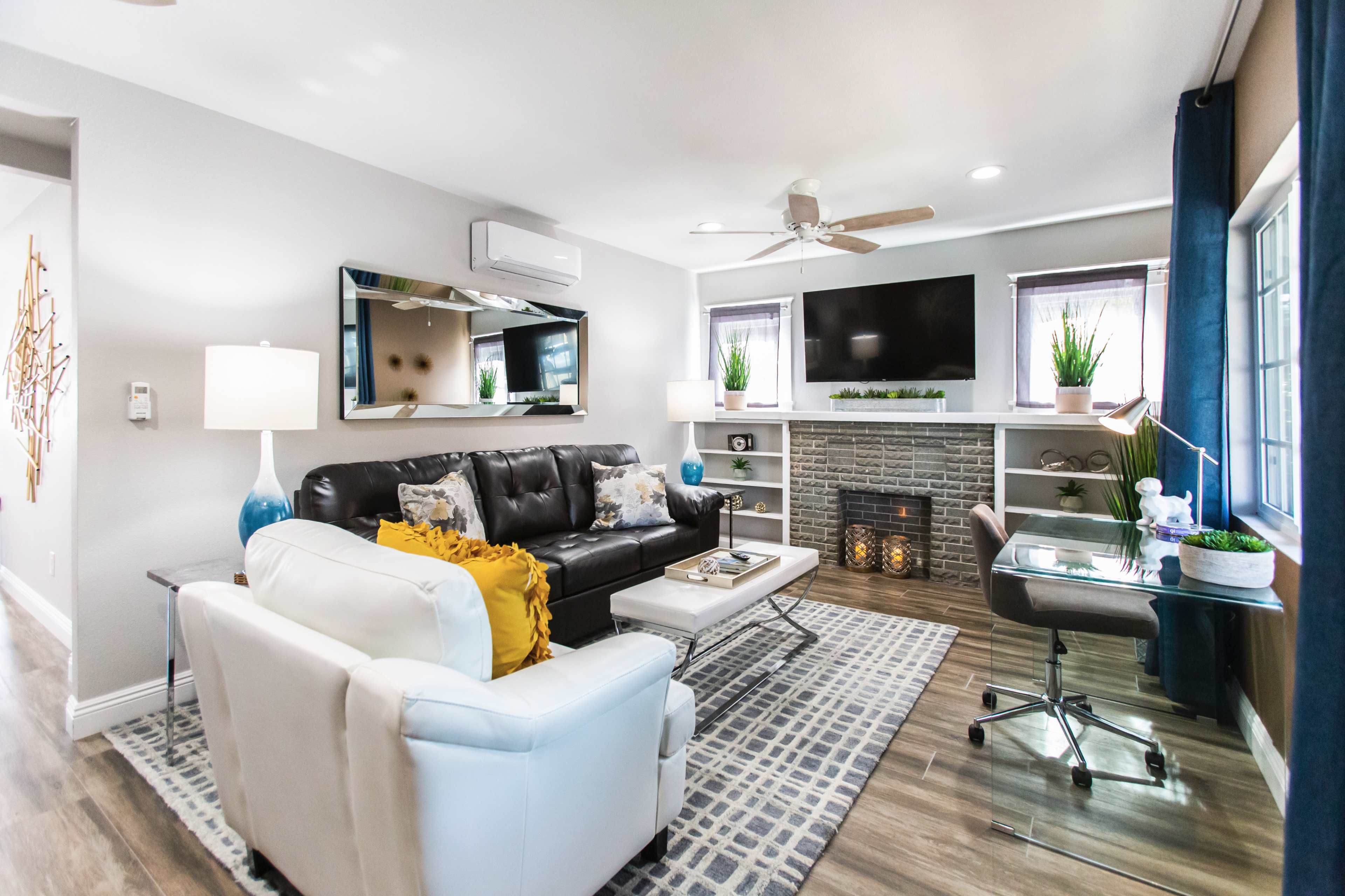 The living room features a black leather sofa, a white armchair, a television mounted above a decorative fireplace, and a desk beside large windows.