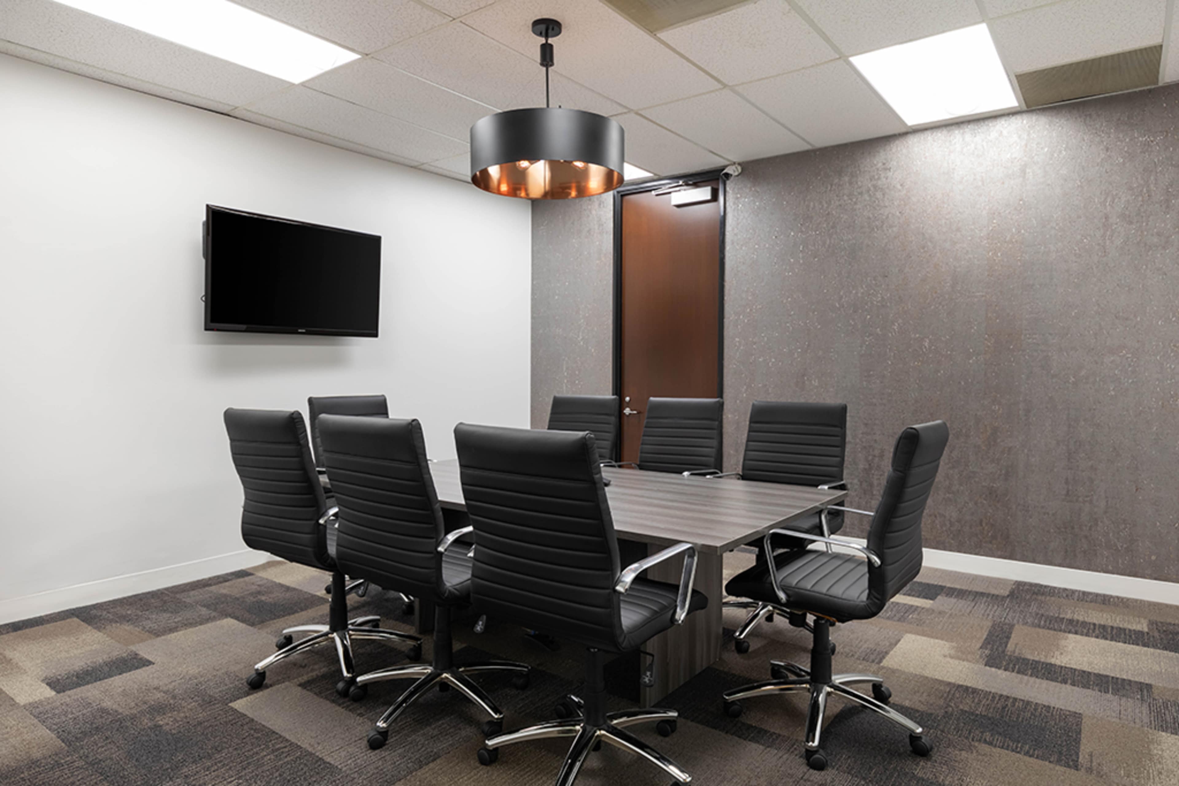 A modern conference room features a rectangular table surrounded by eight black leather chairs, with a television mounted on the wall and a pendant light overhead.