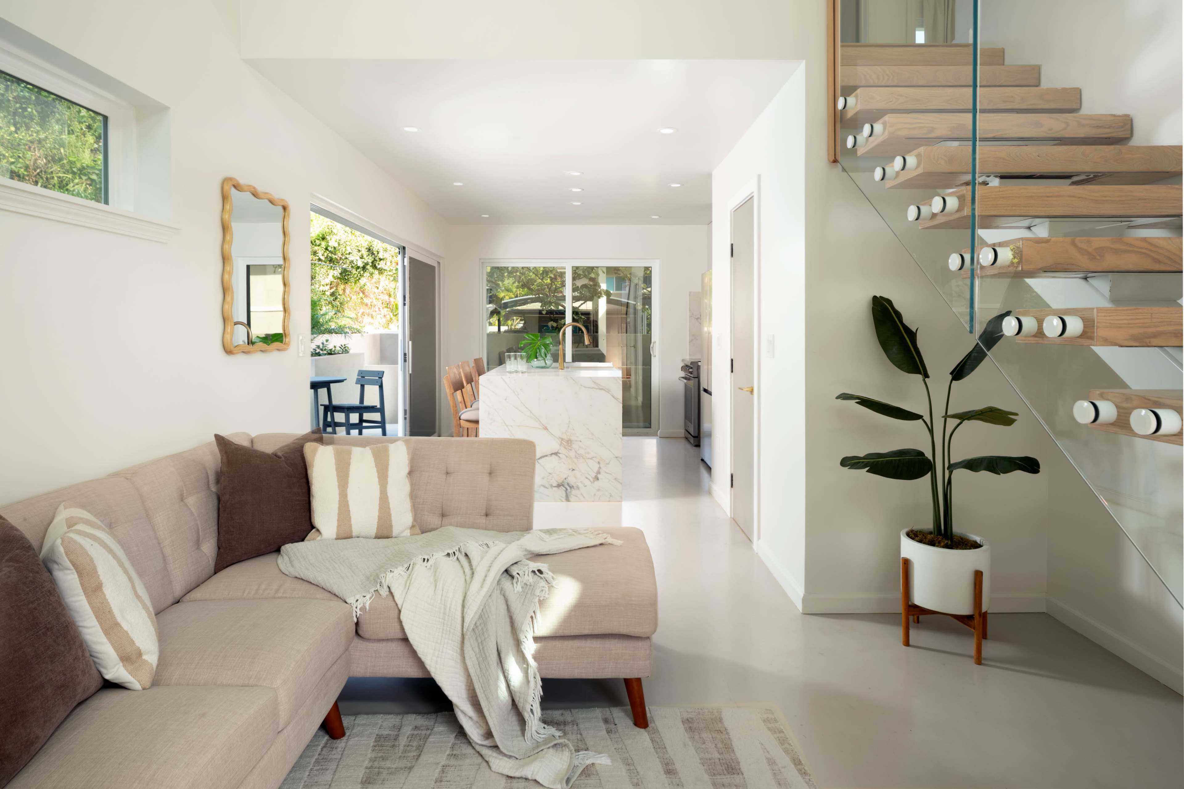 The image shows a bright and modern living space featuring a light-colored sectional sofa, a small dining area with stools, and a staircase with wooden steps and glass railings.