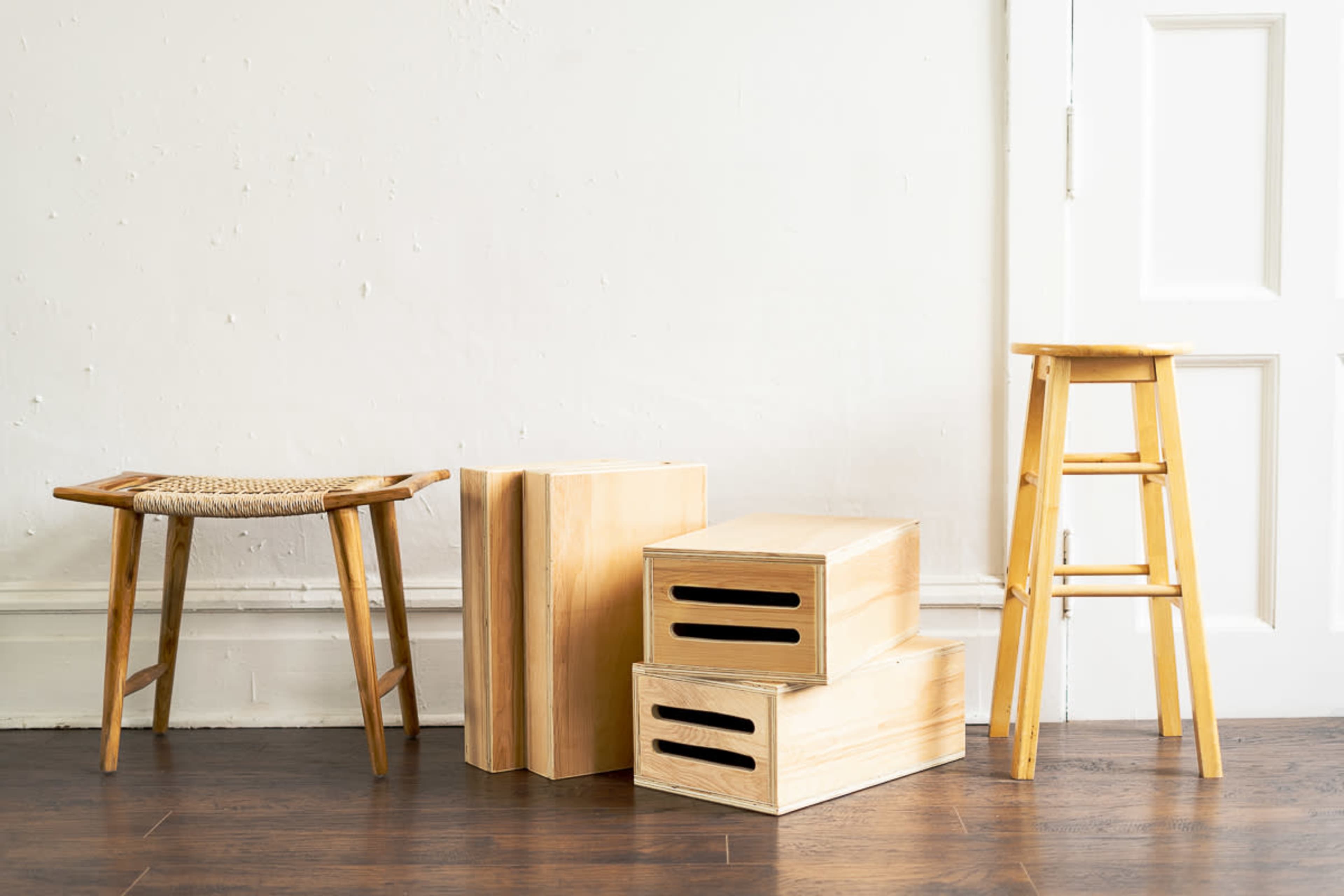 Various wooden furniture pieces, including a woven stool, stacked wooden boxes, and a tall wooden stool, set against a plain white wall.