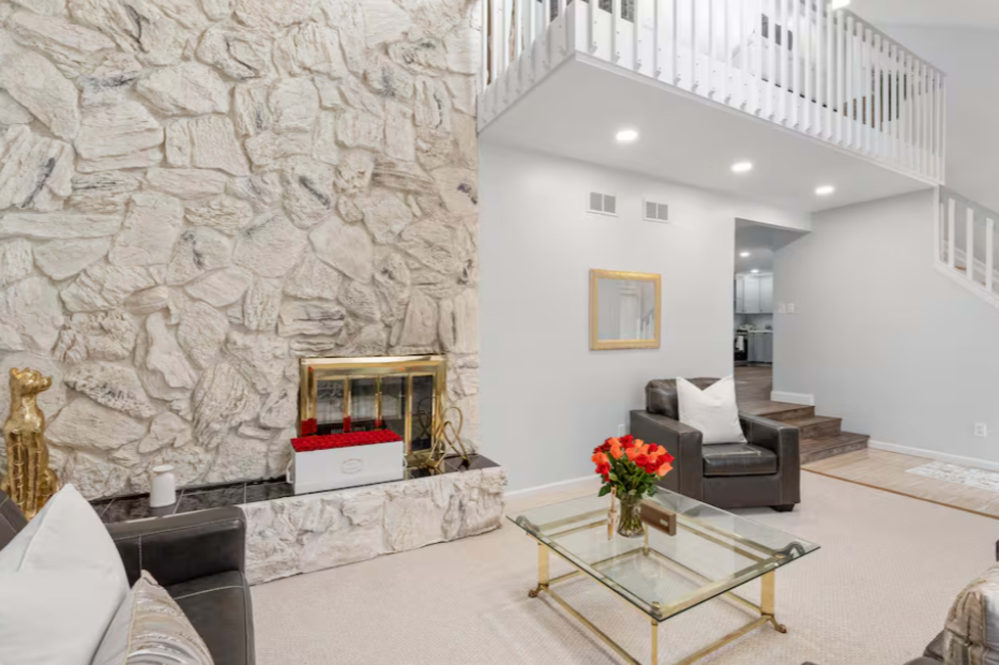 The image shows a living room featuring a stone fireplace, a glass coffee table, and a staircase leading to an upper level.