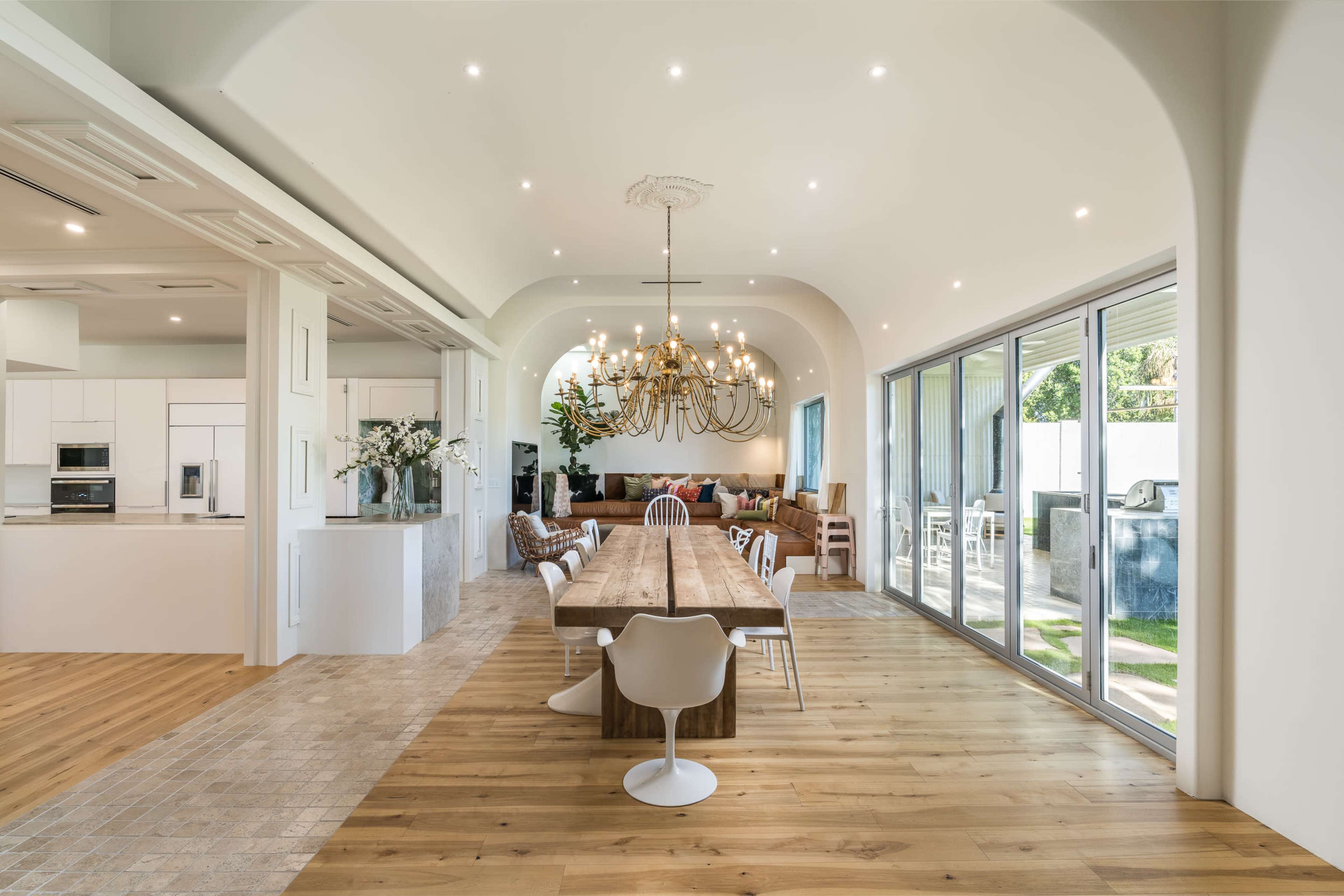 A modern dining area with a long wooden table surrounded by white chairs, a large chandelier overhead, and large glass doors leading to an outdoor space.