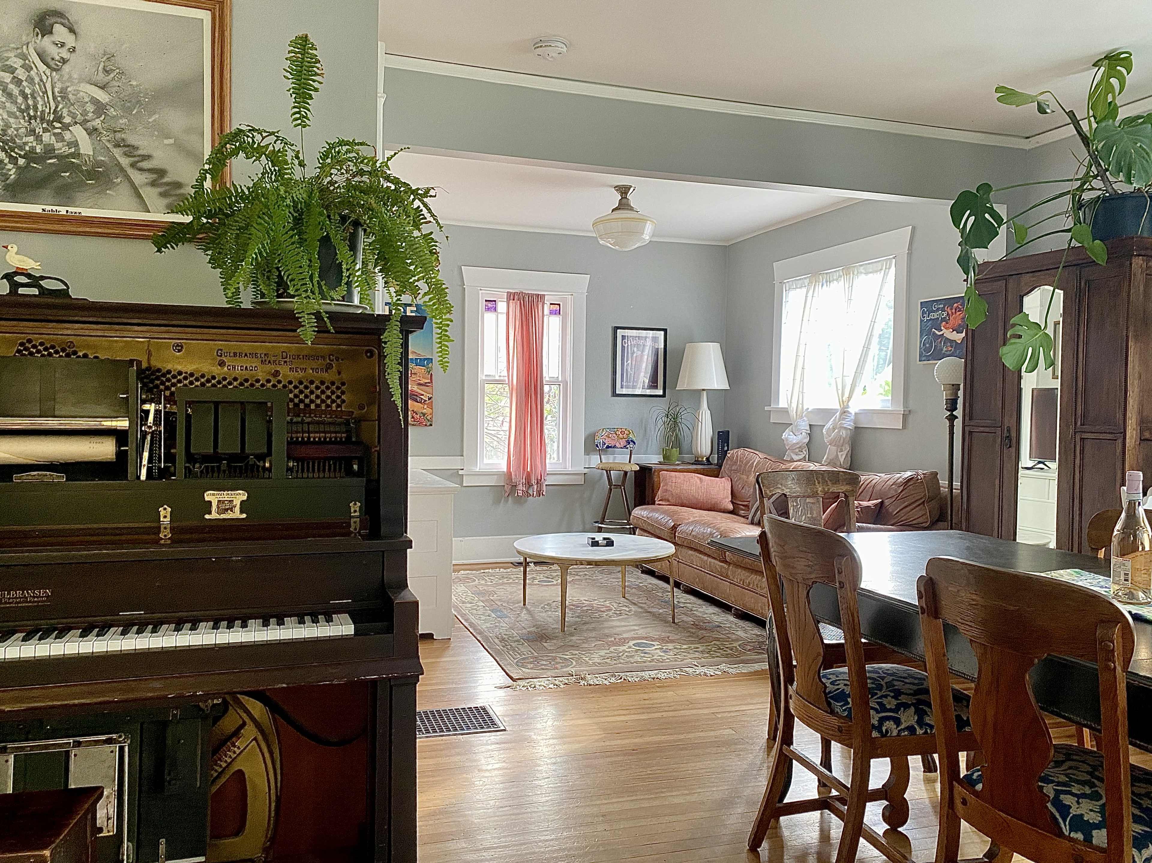 The image shows a cozy living space with a vintage piano, a dining table with chairs, a leather couch, and various plants scattered throughout the room.