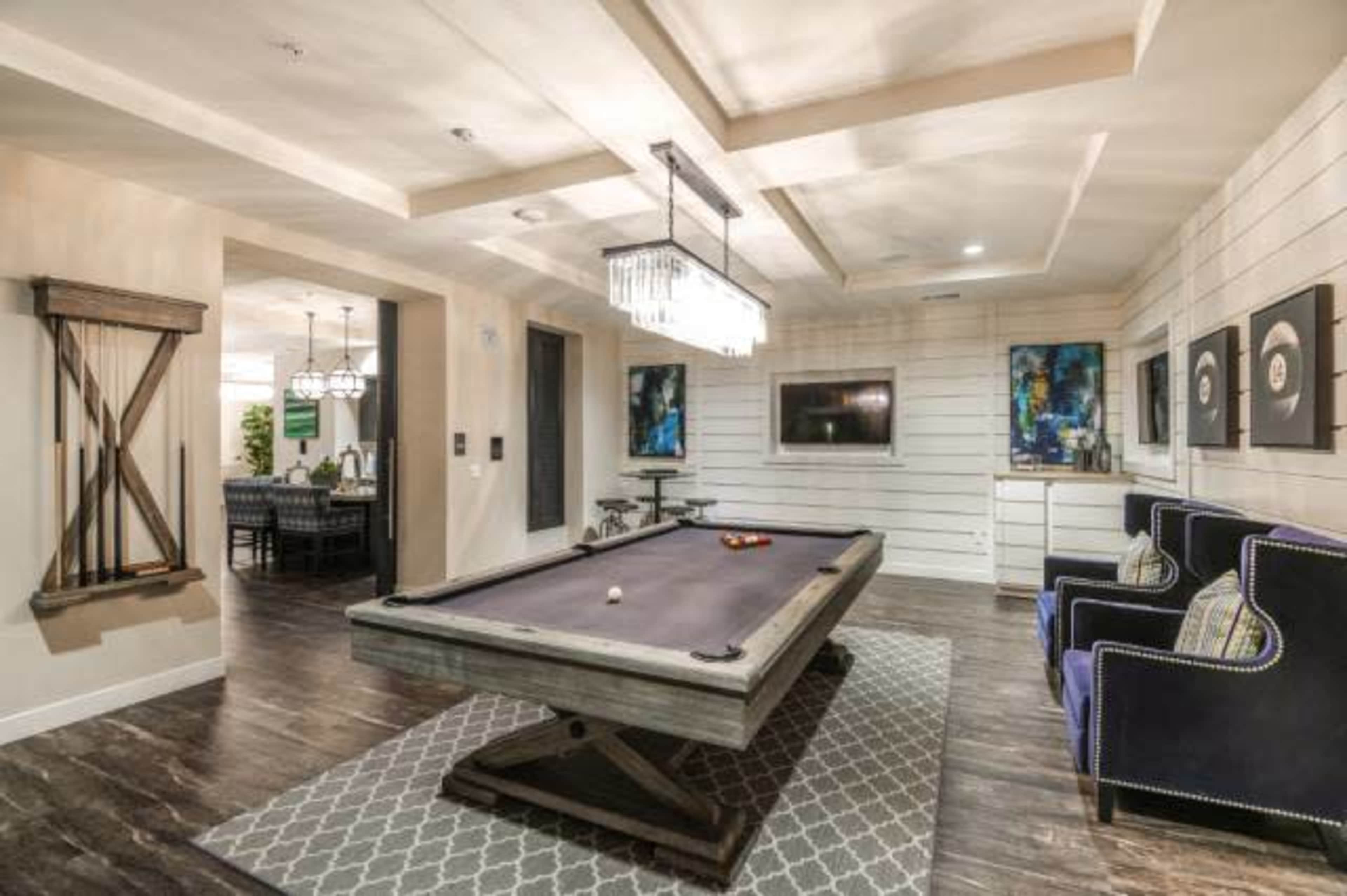 The image features a game room with a pool table, two armchairs, and a modern chandelier, set against a backdrop of paneled walls and artwork.