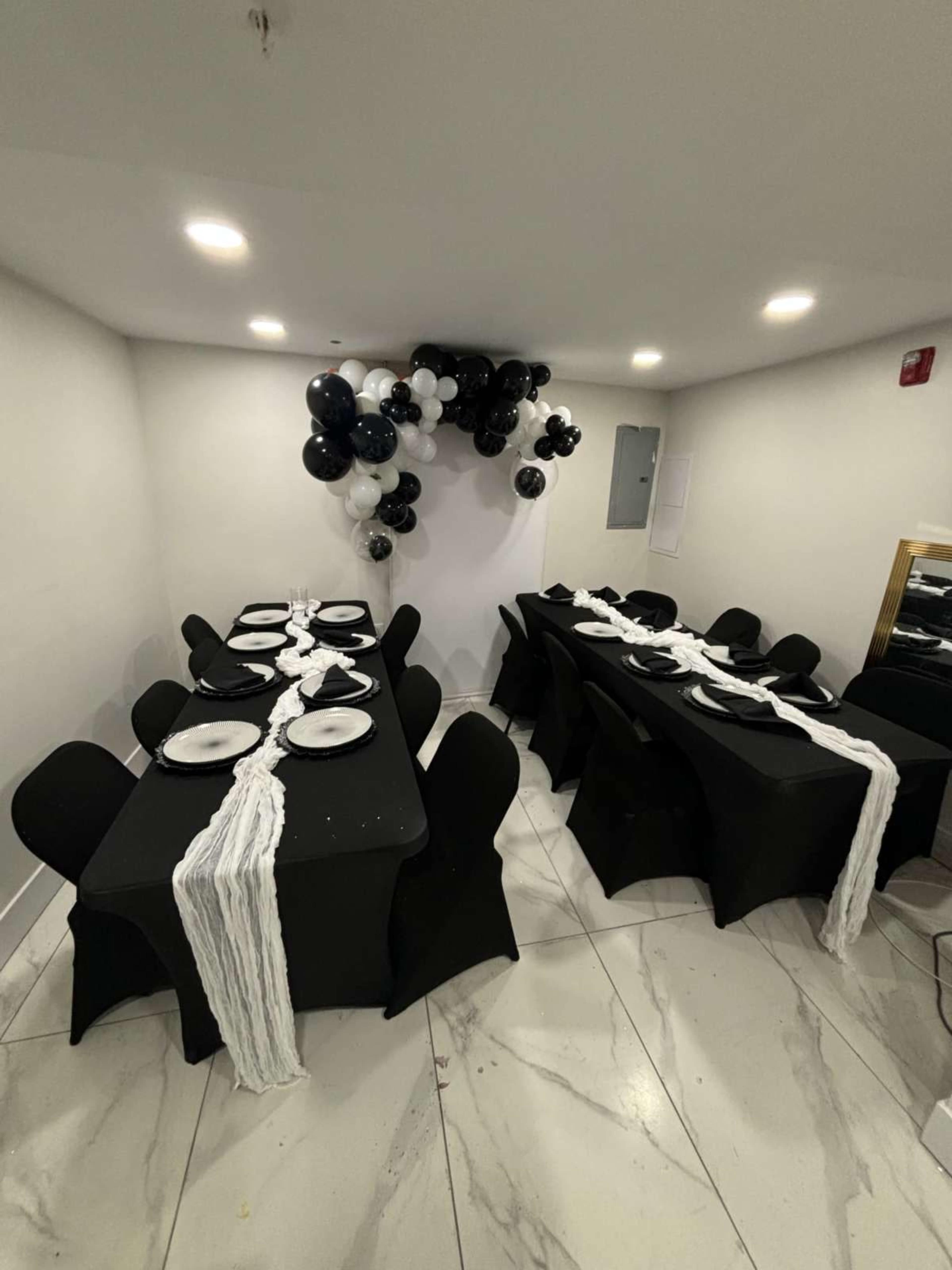 The image shows a room set up for a formal event, featuring two tables covered with black tablecloths, each adorned with white plates and a white runner, and decorated with a cluster of black and white balloons above.
