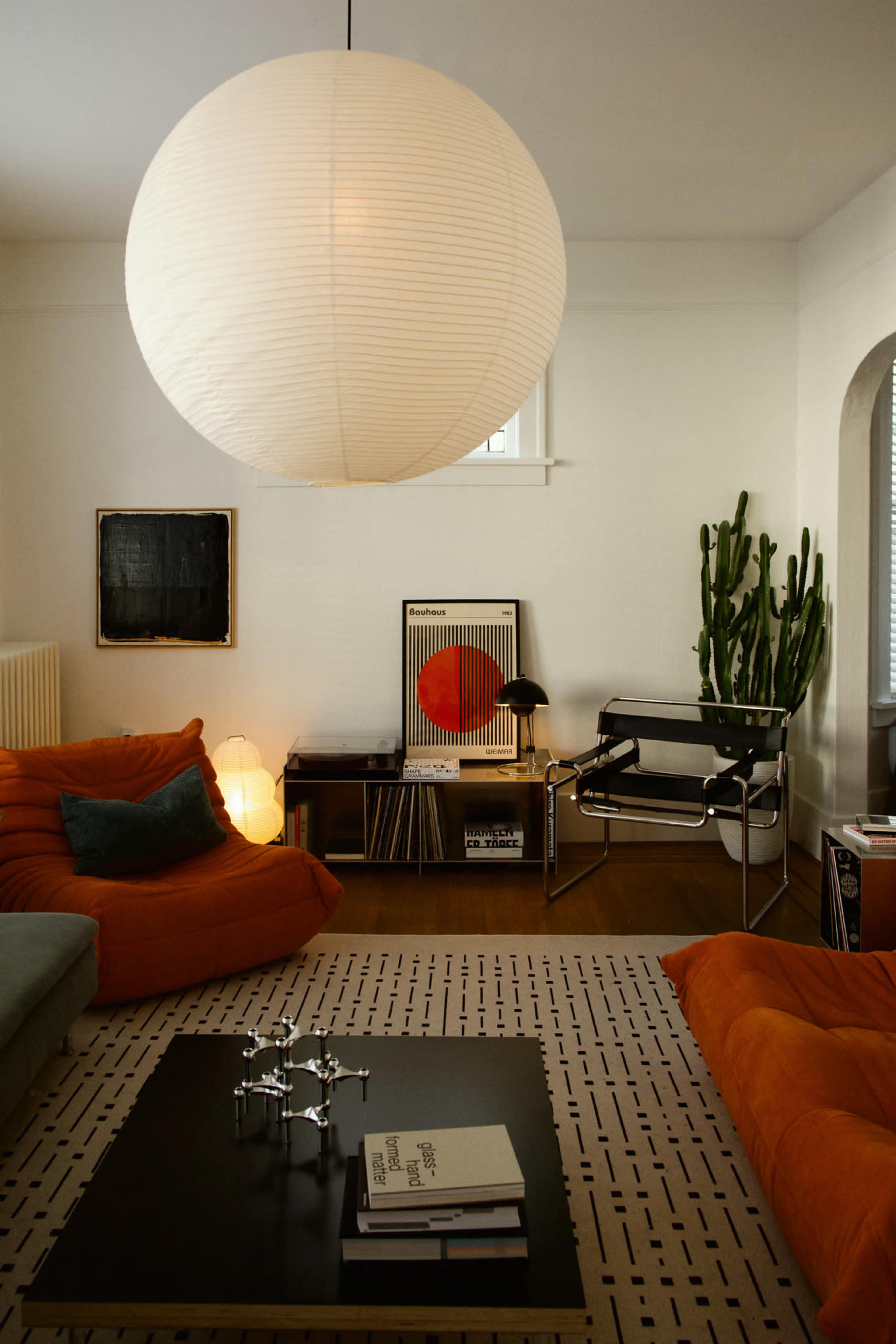 The room features a modern, minimalist design with two orange lounge chairs, a black coffee table, a large paper lantern pendant light, and various art pieces on the walls.