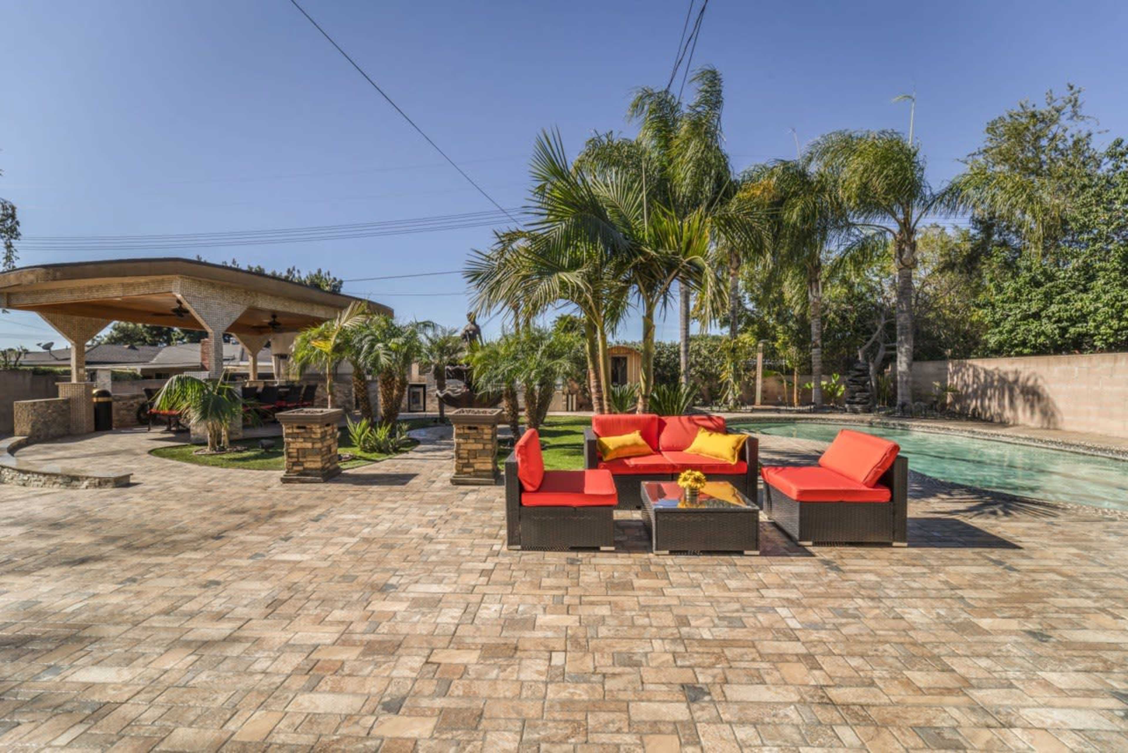 A backyard with a stone patio, a pool, palm trees, and a seating area featuring red and yellow cushions.