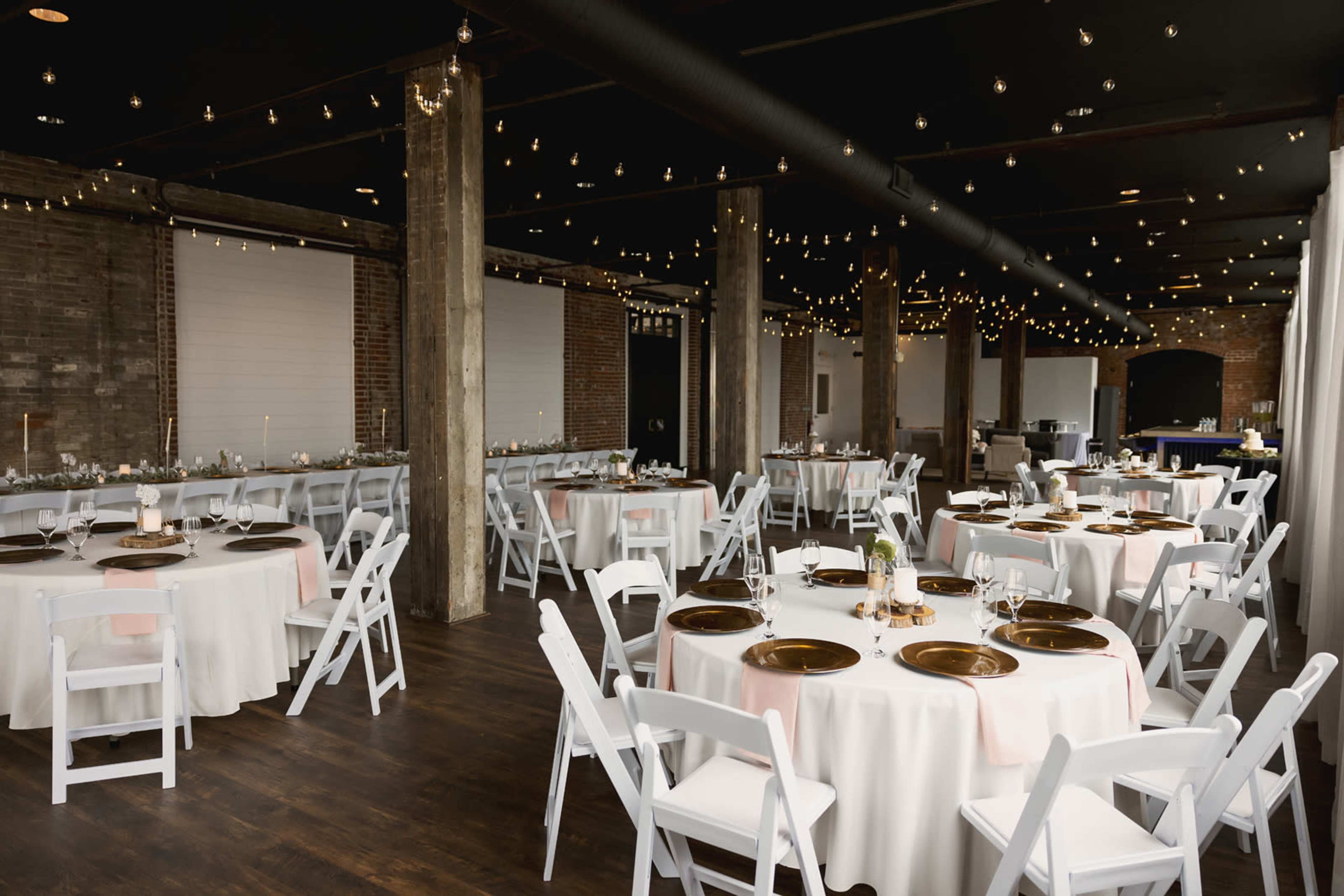 A spacious event venue with round tables set for a formal dinner, surrounded by string lights and exposed brick walls.
