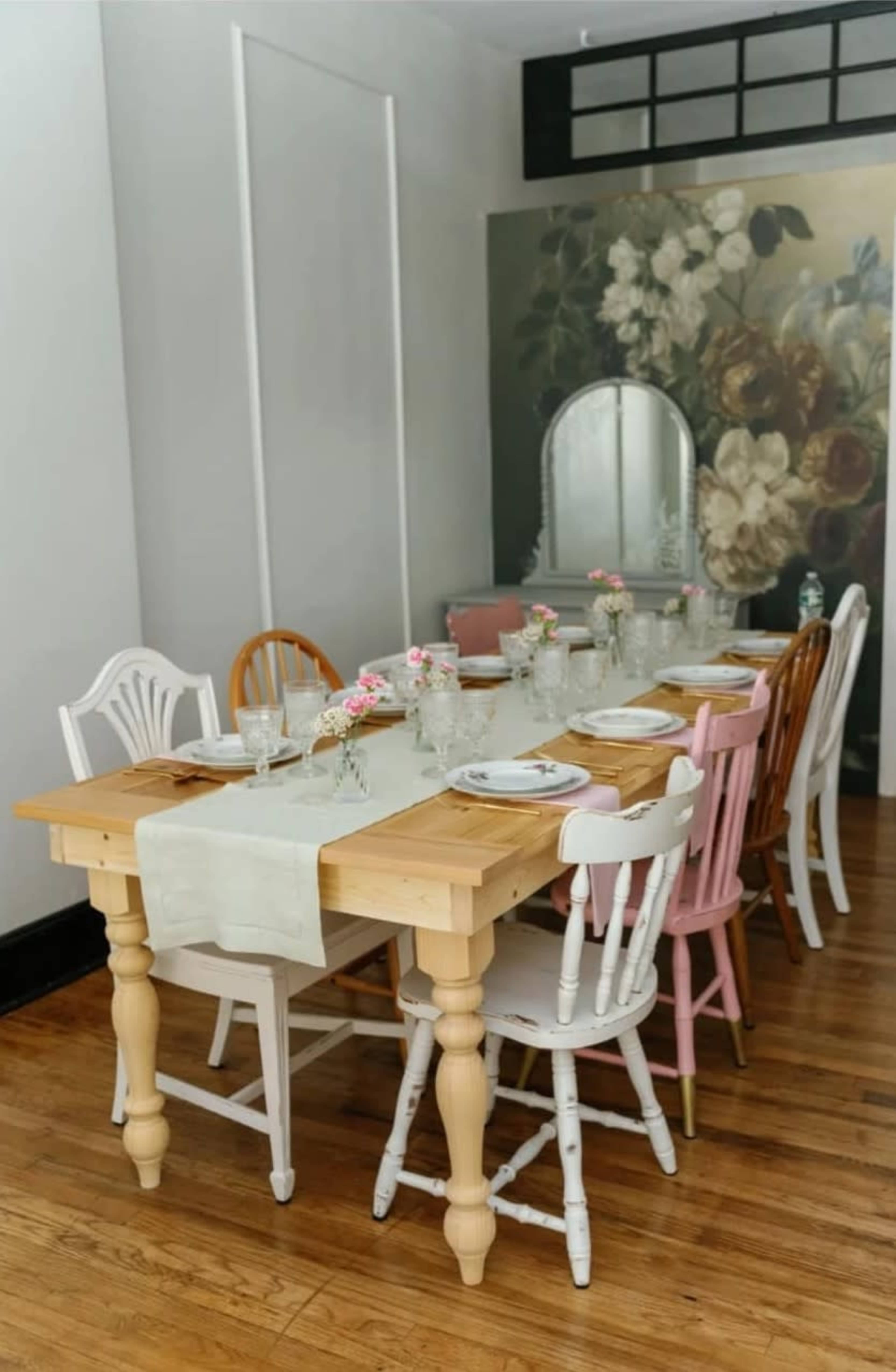 A long wooden dining table is set with plates and glasses, surrounded by a mix of pastel-colored chairs in a room with floral wallpaper and a decorative mirror.