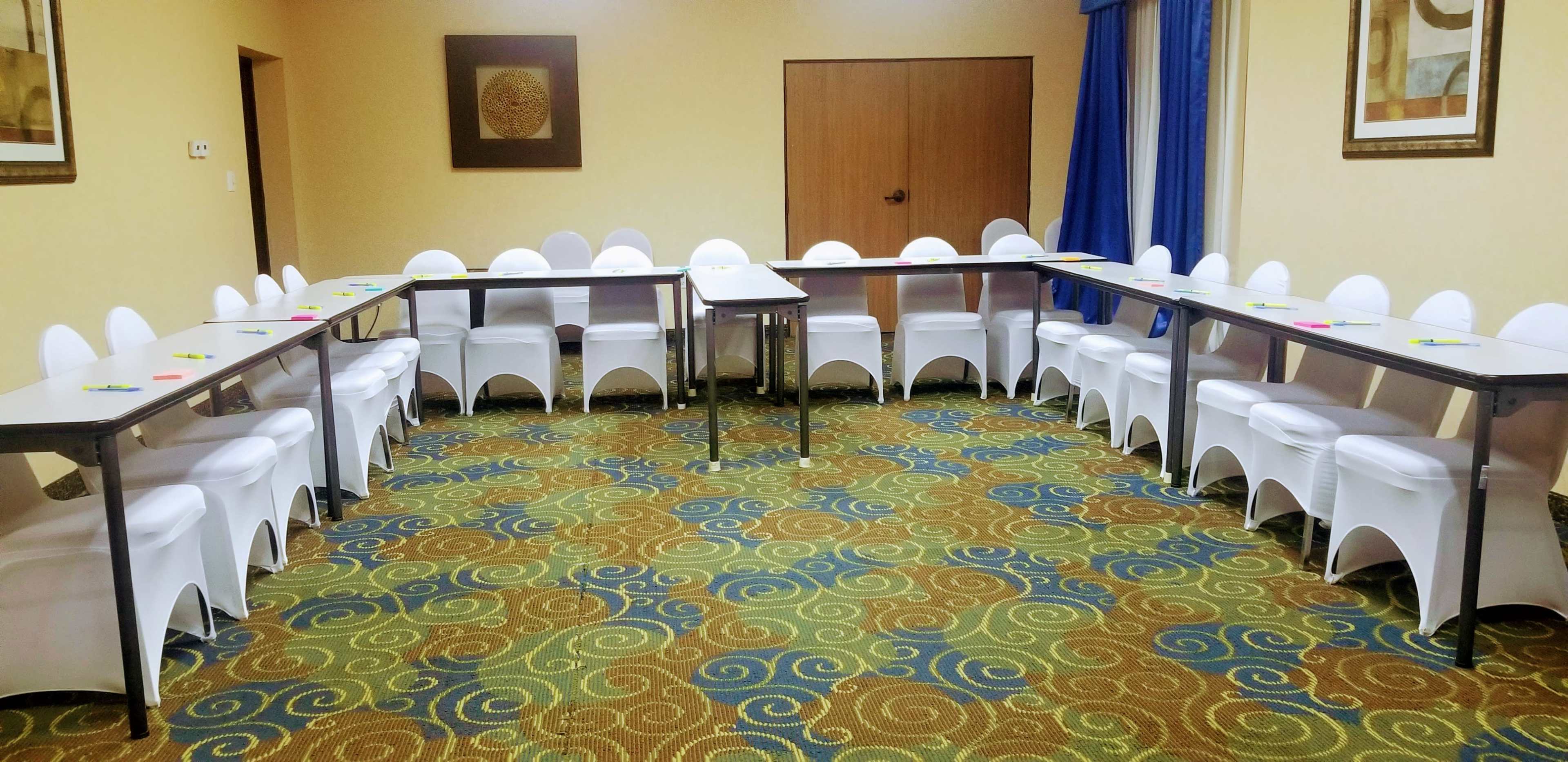 A meeting room features multiple tables arranged in a U-shape with white chairs around them, set on a patterned carpet.