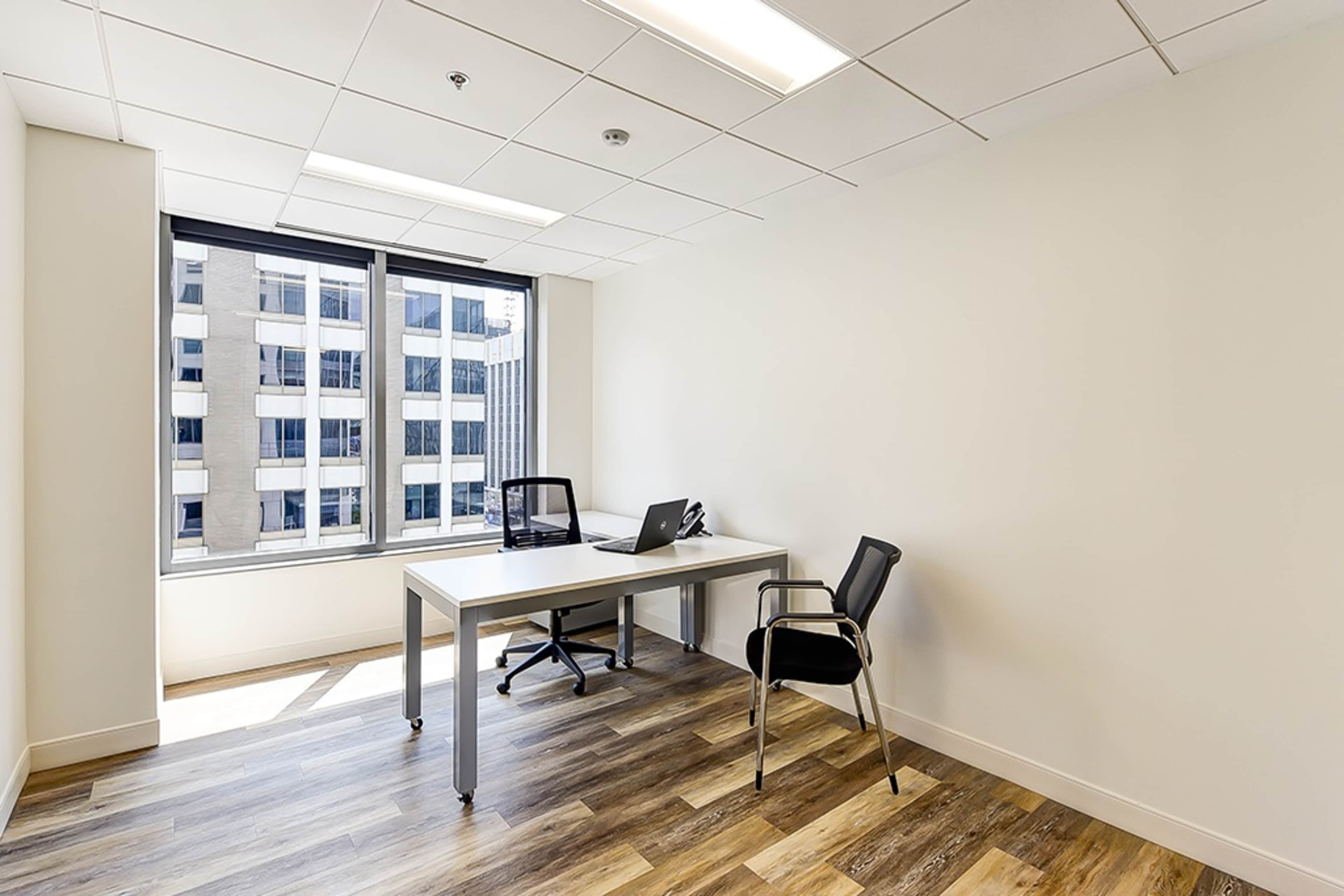 A bright, minimalistic office space features a desk with a laptop and an empty chair by a large window overlooking a cityscape.