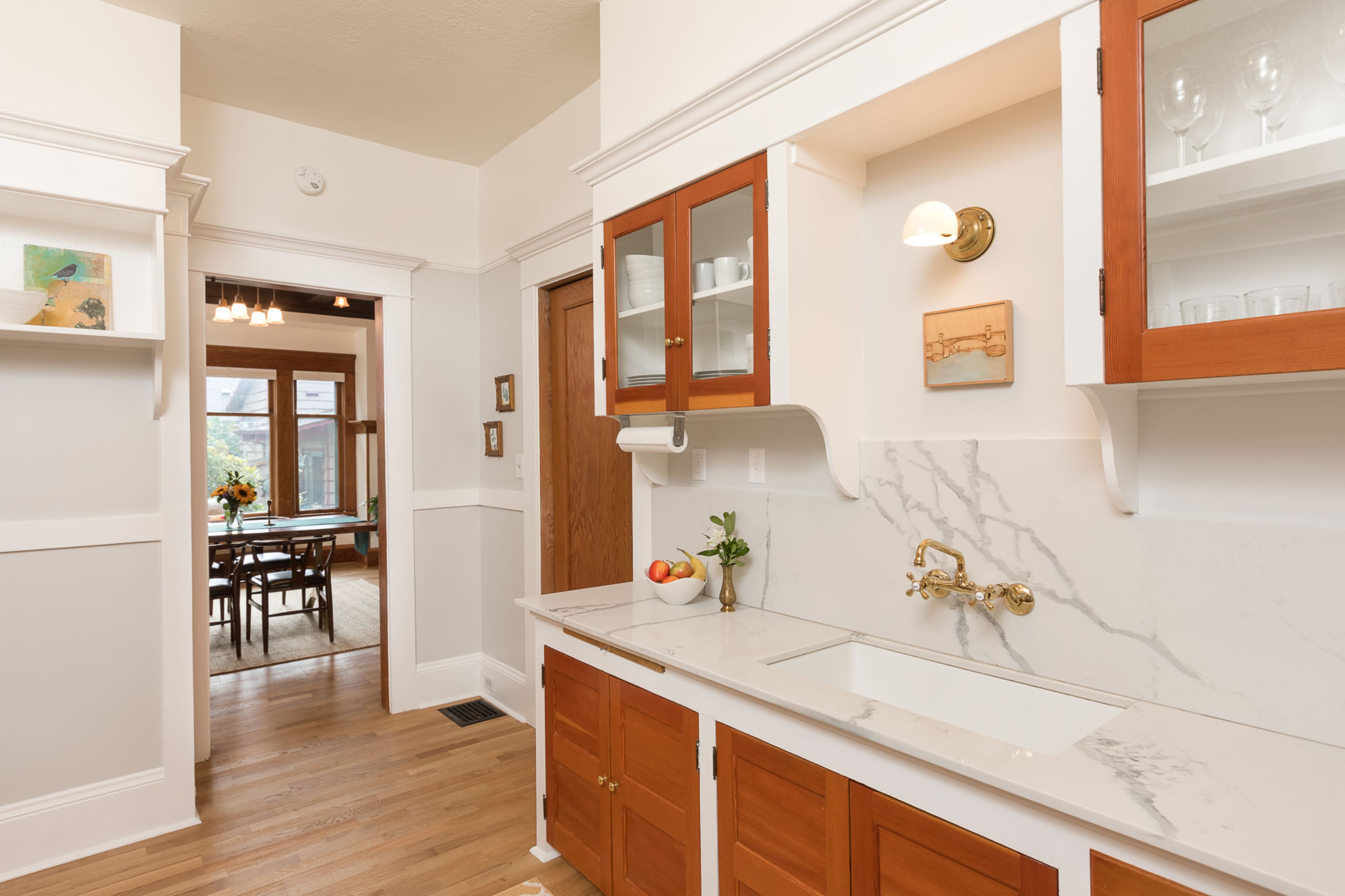 A bright kitchen features a marble countertop with a farmhouse sink and wooden cabinets, leading to a dining area visible in the background.