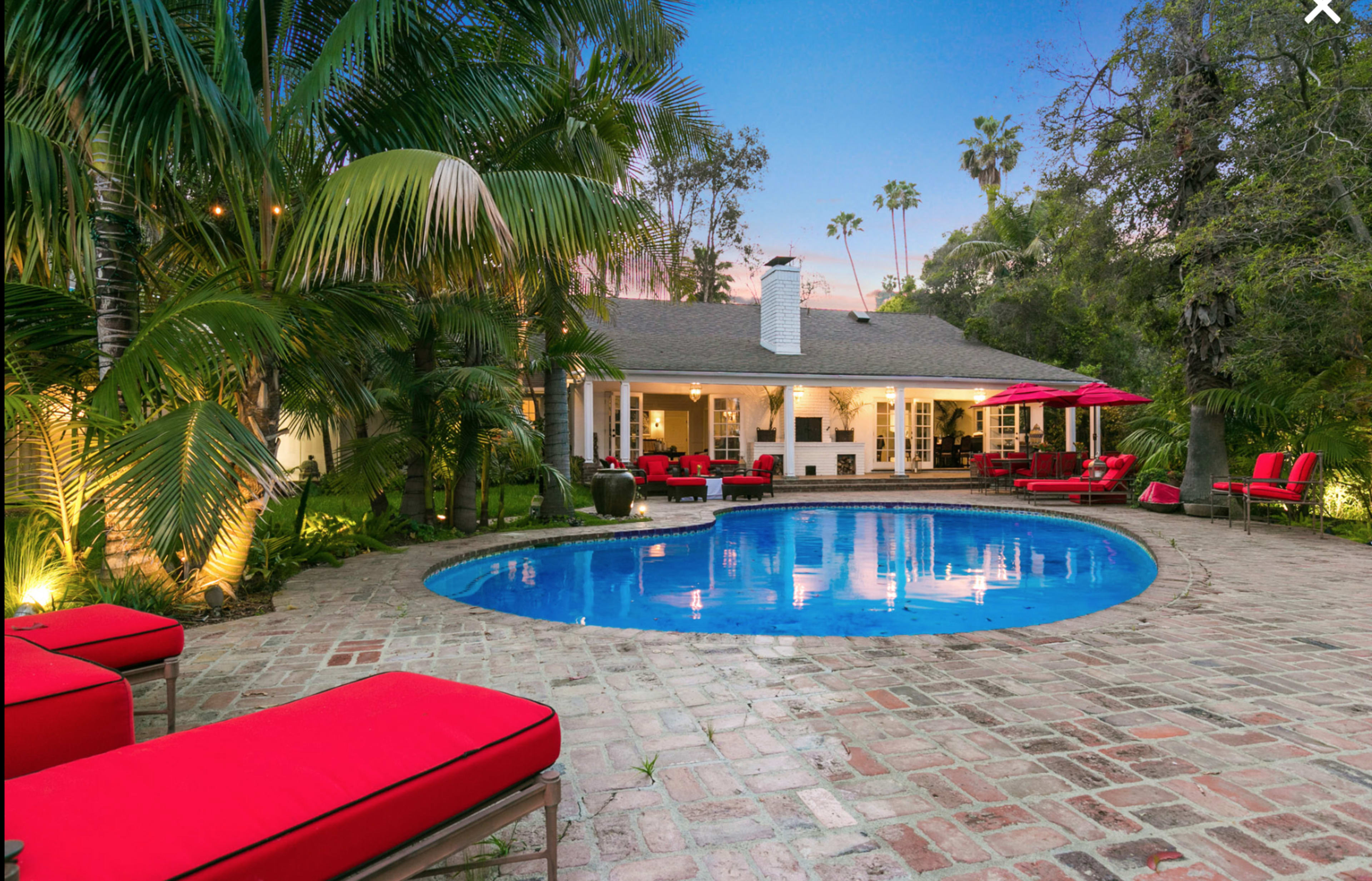 The image shows a backyard with a swimming pool surrounded by lush greenery and red lounge chairs, with a single-story house in the background.