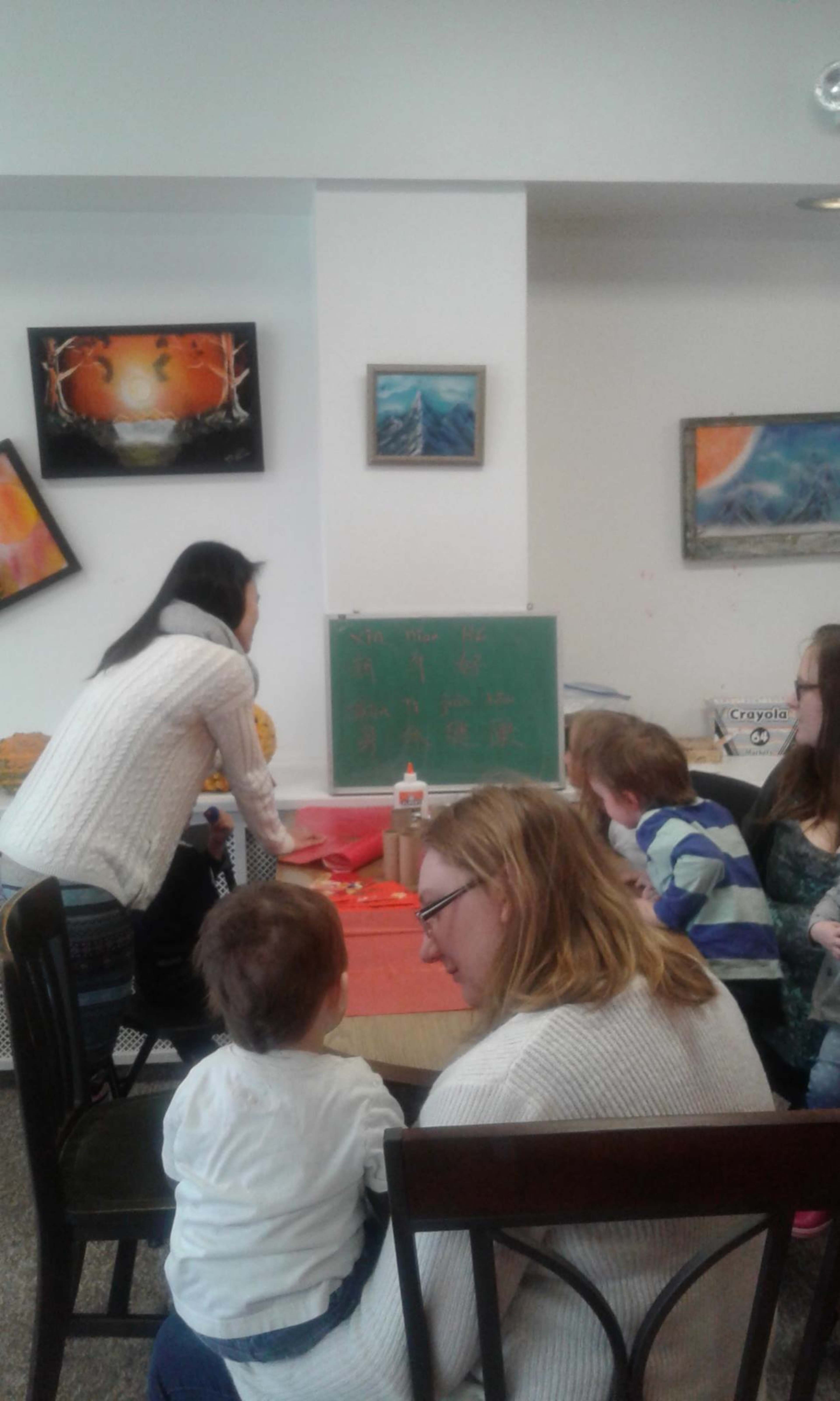 A group of children and adults gather around a table where a woman is standing next to a chalkboard, engaging in an activity in a brightly decorated room.