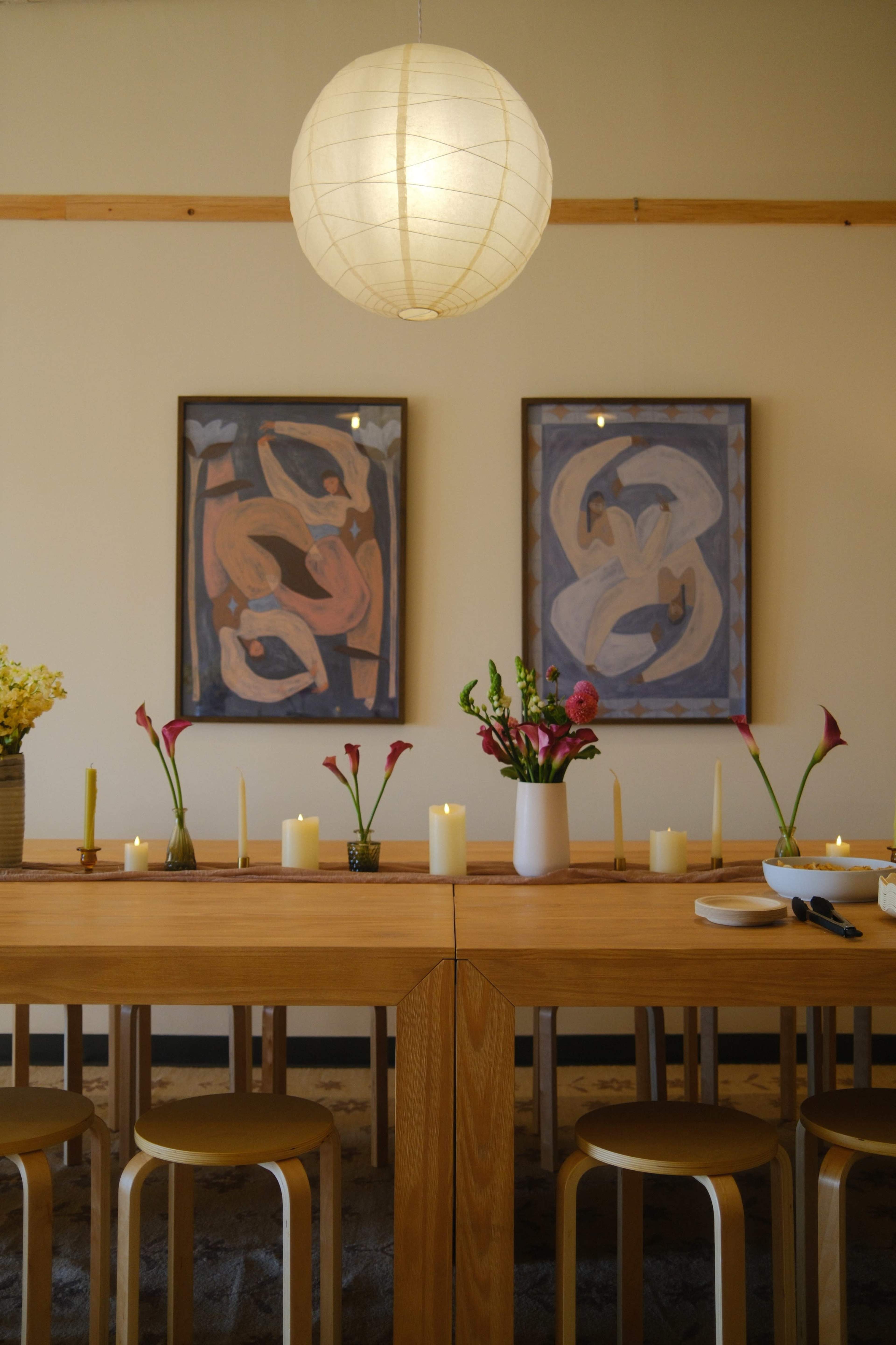 A wooden dining table is set with candles and a vase of flowers, flanked by two abstract paintings on the walls and illuminated by a large paper lantern.
