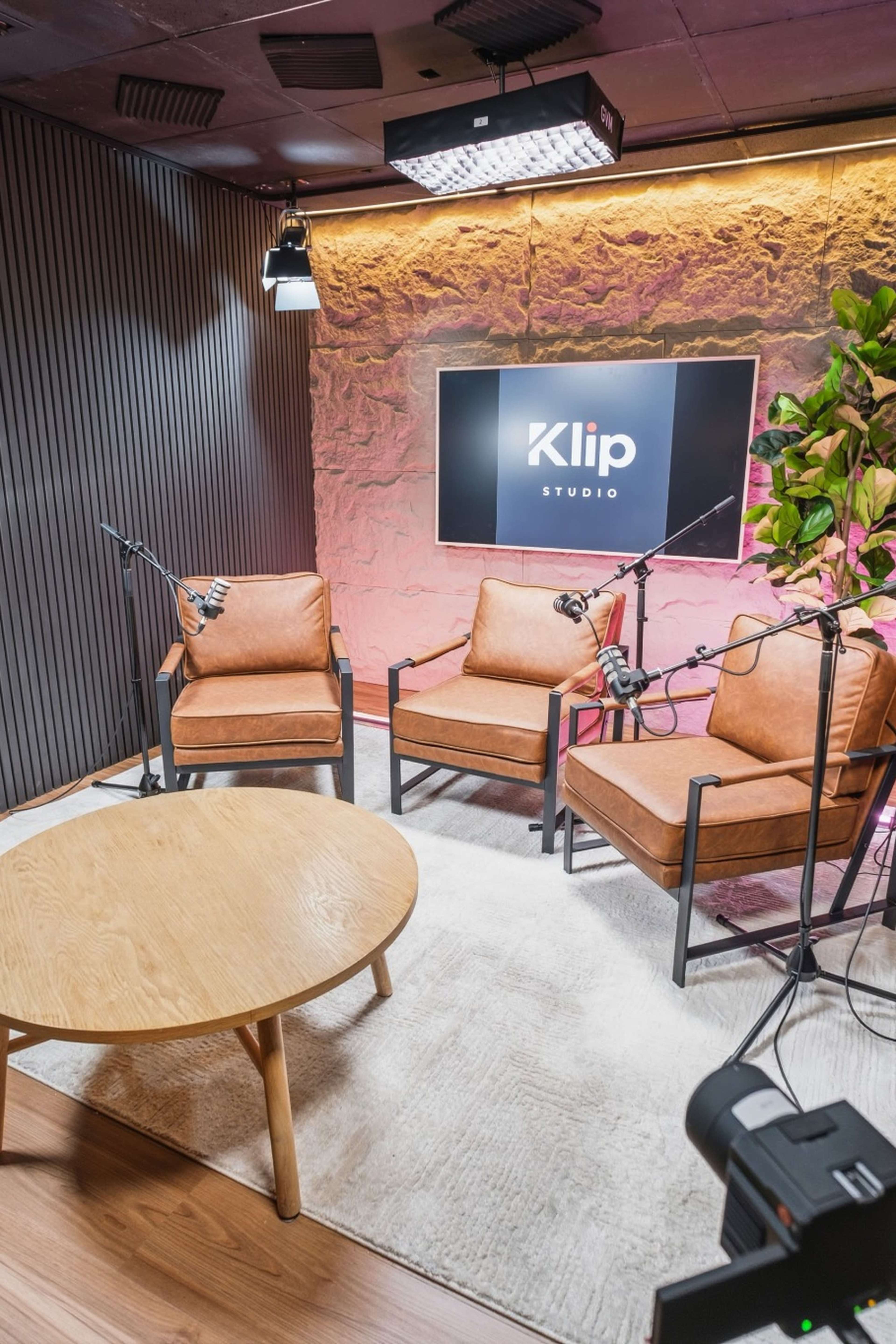 The image shows a small studio space arranged with three brown armchairs around a circular wooden table, with a screen displaying "Klip Studio" in the background.