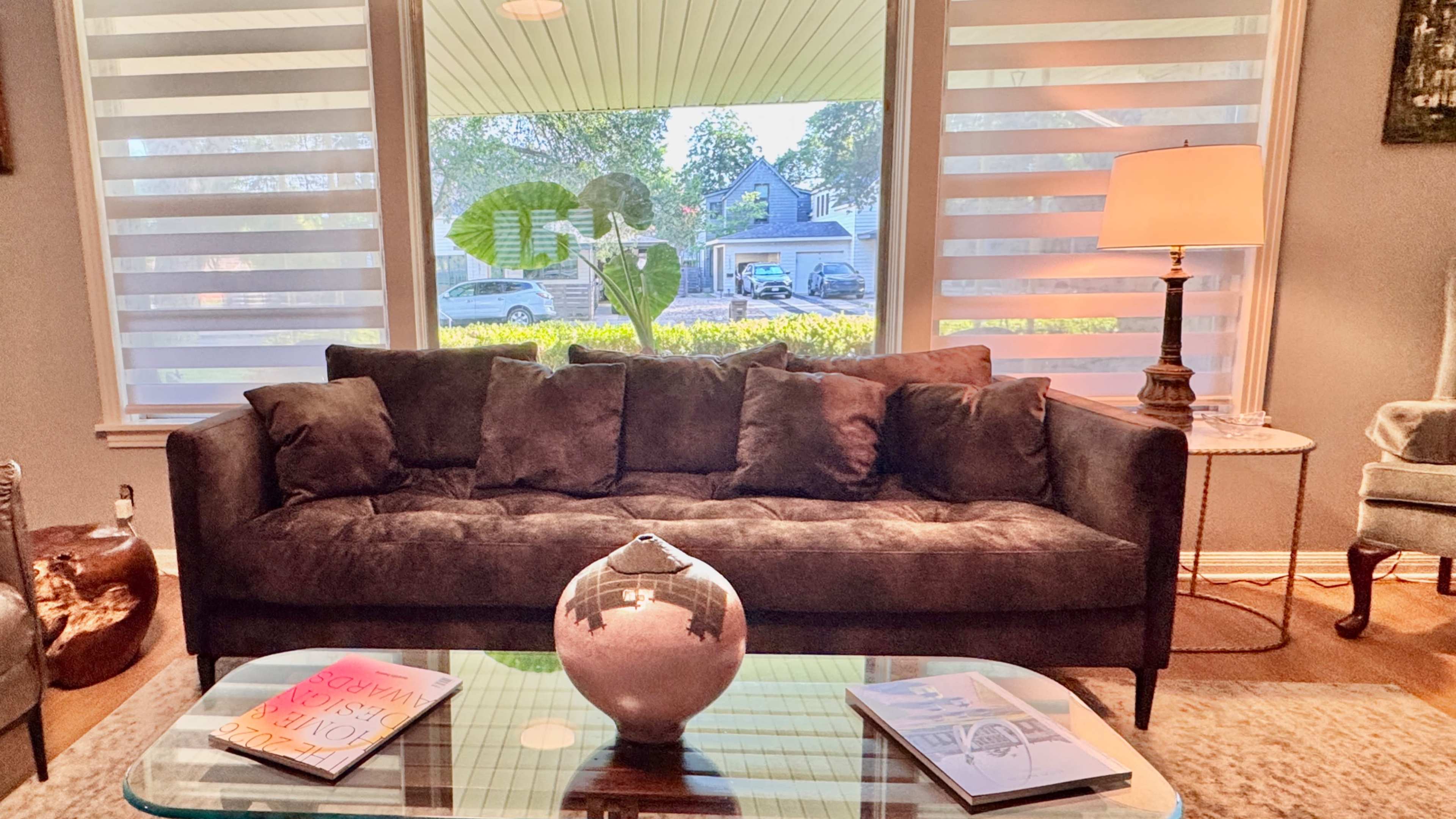 The scene features a cozy living room with a brown sofa facing a large window, a glass coffee table adorned with magazines and a decorative vase, and a lamp providing light next to the sofa.