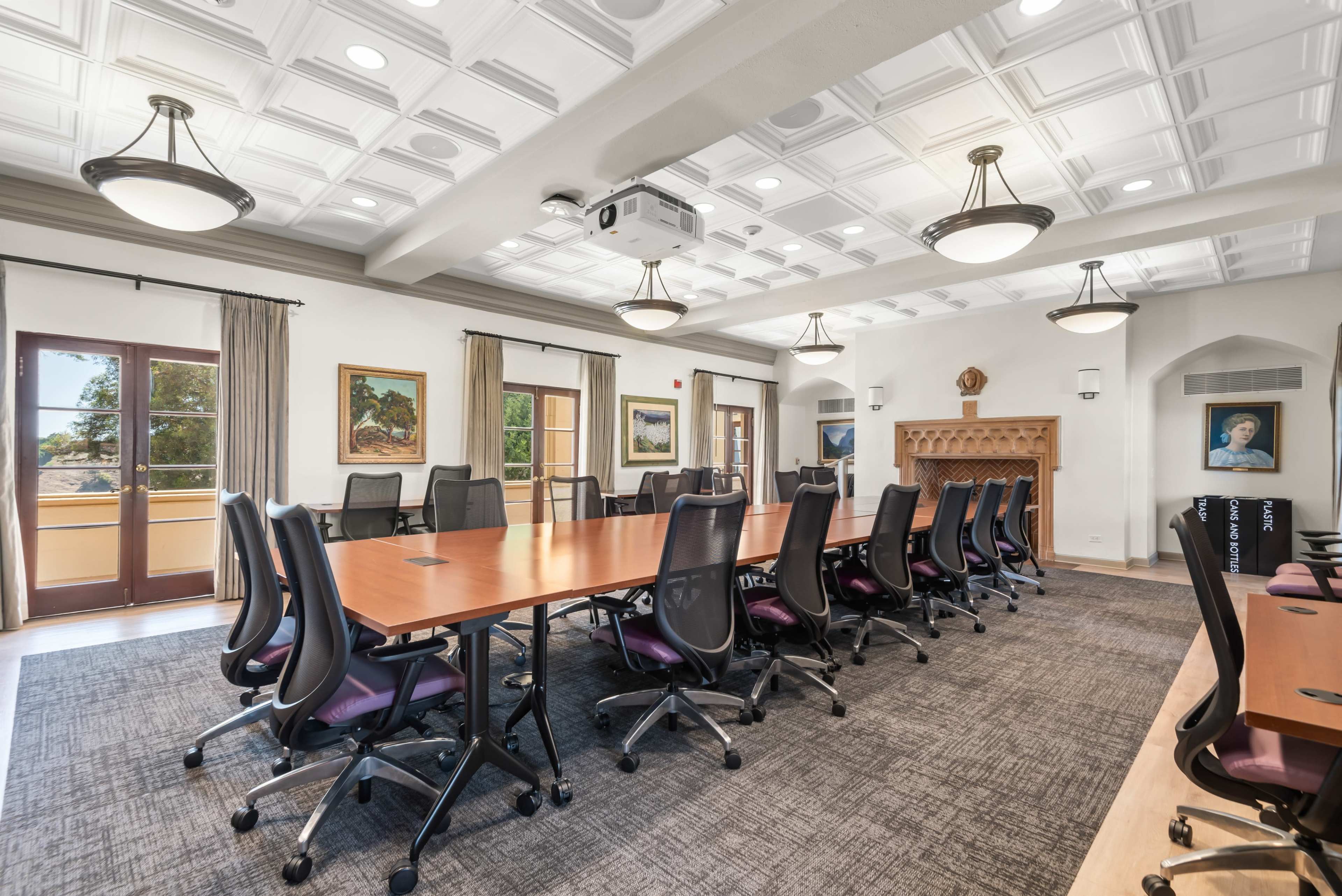 The image shows a spacious conference room featuring a long wooden table surrounded by ergonomic chairs, with large windows providing natural light and a decorative fireplace.