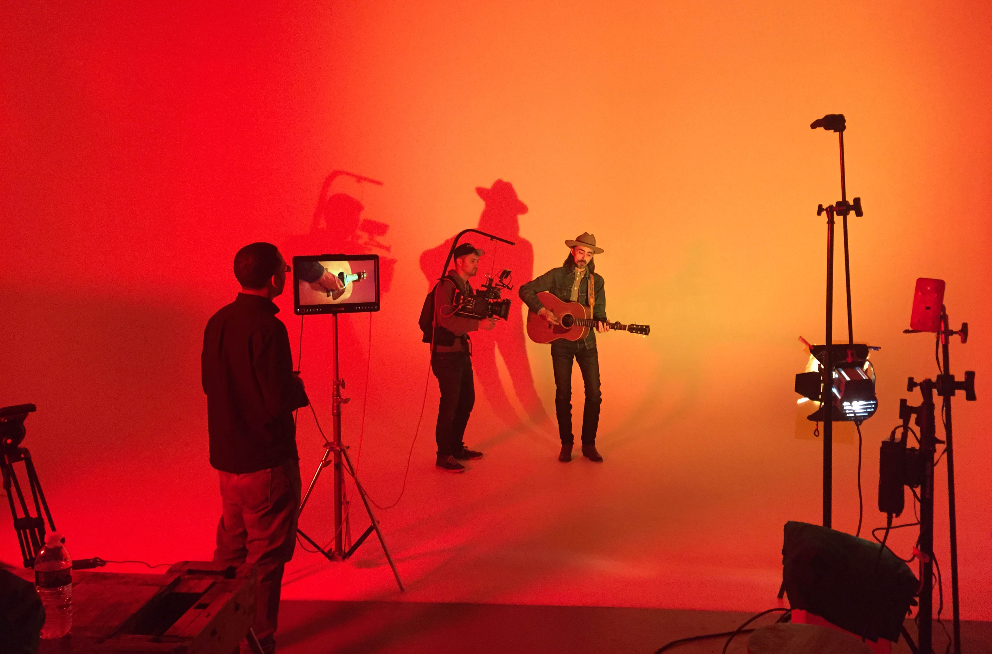 A musician stands with a guitar against a vibrant orange backdrop while two crew members operate cameras and lighting equipment.