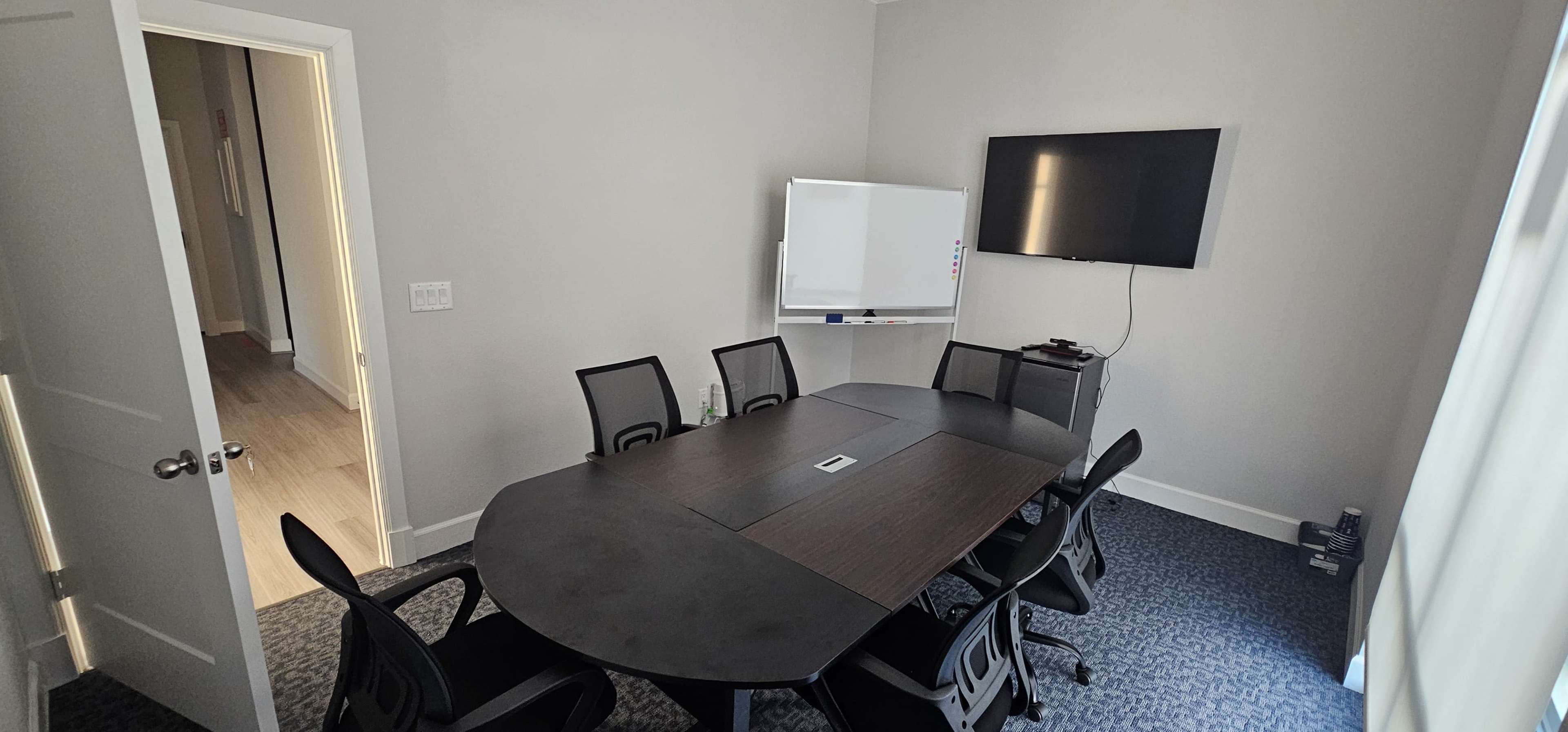 A modern meeting room features a long, oval conference table surrounded by ergonomic chairs, with a whiteboard and a wall-mounted television present.