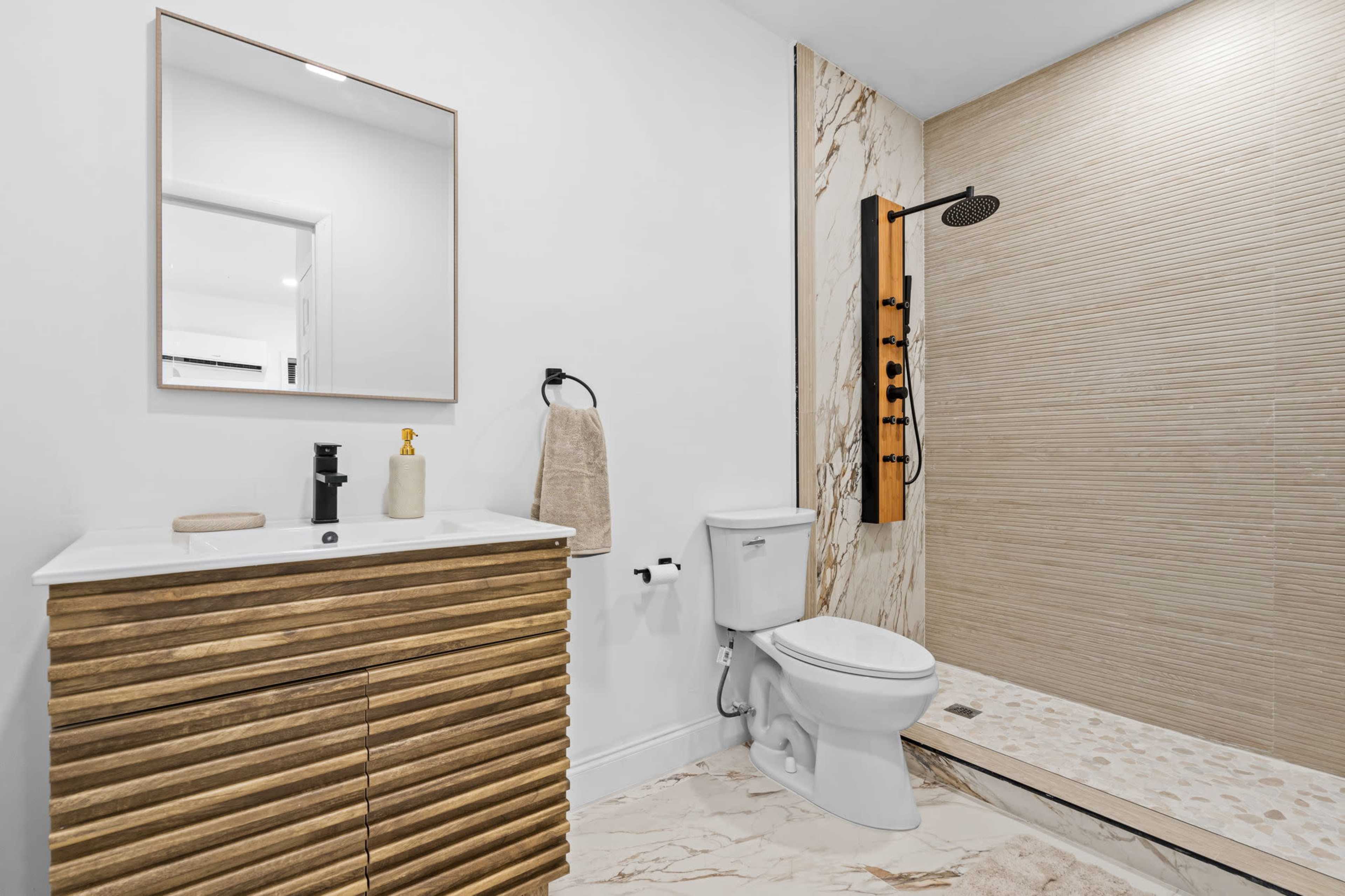 The image shows a modern bathroom featuring a wooden vanity with a sink, a toilet, and a shower area with tile and stone finishes.