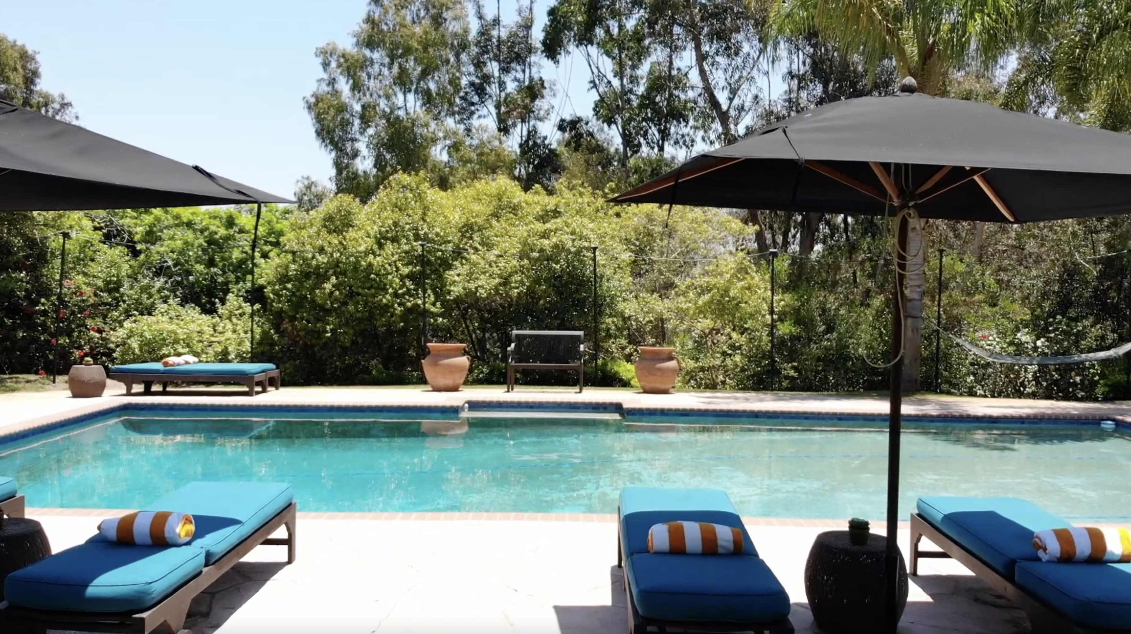 A rectangular swimming pool is surrounded by lounge chairs, umbrellas, and lush greenery.