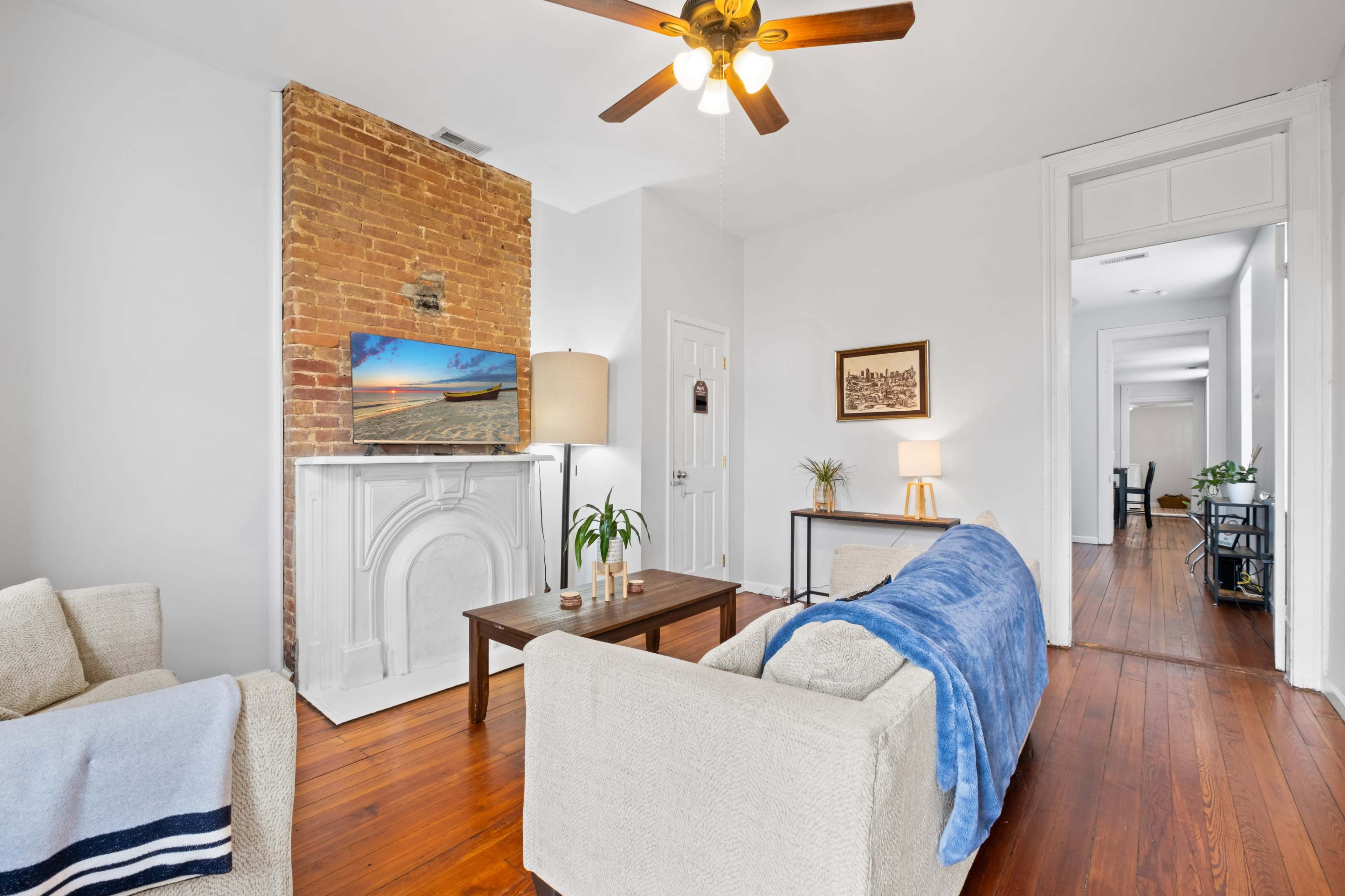 The living room features a brick wall, a television mounted above a white mantel, and two sofas with a blue throw blanket on one.