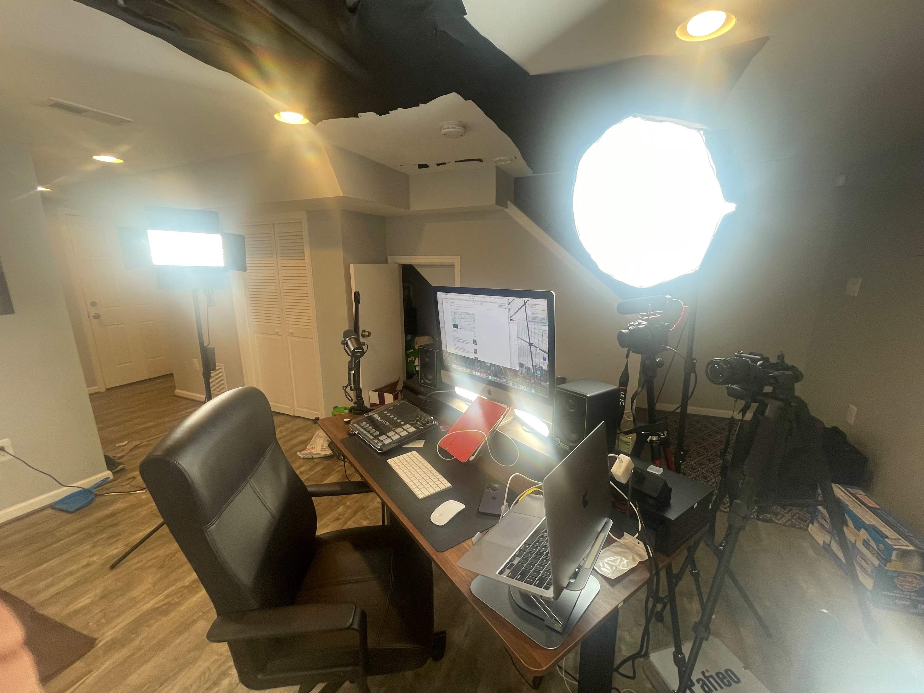 A well-equipped home office setup featuring a desk with multiple computers, cameras, and lighting equipment in a basement environment.