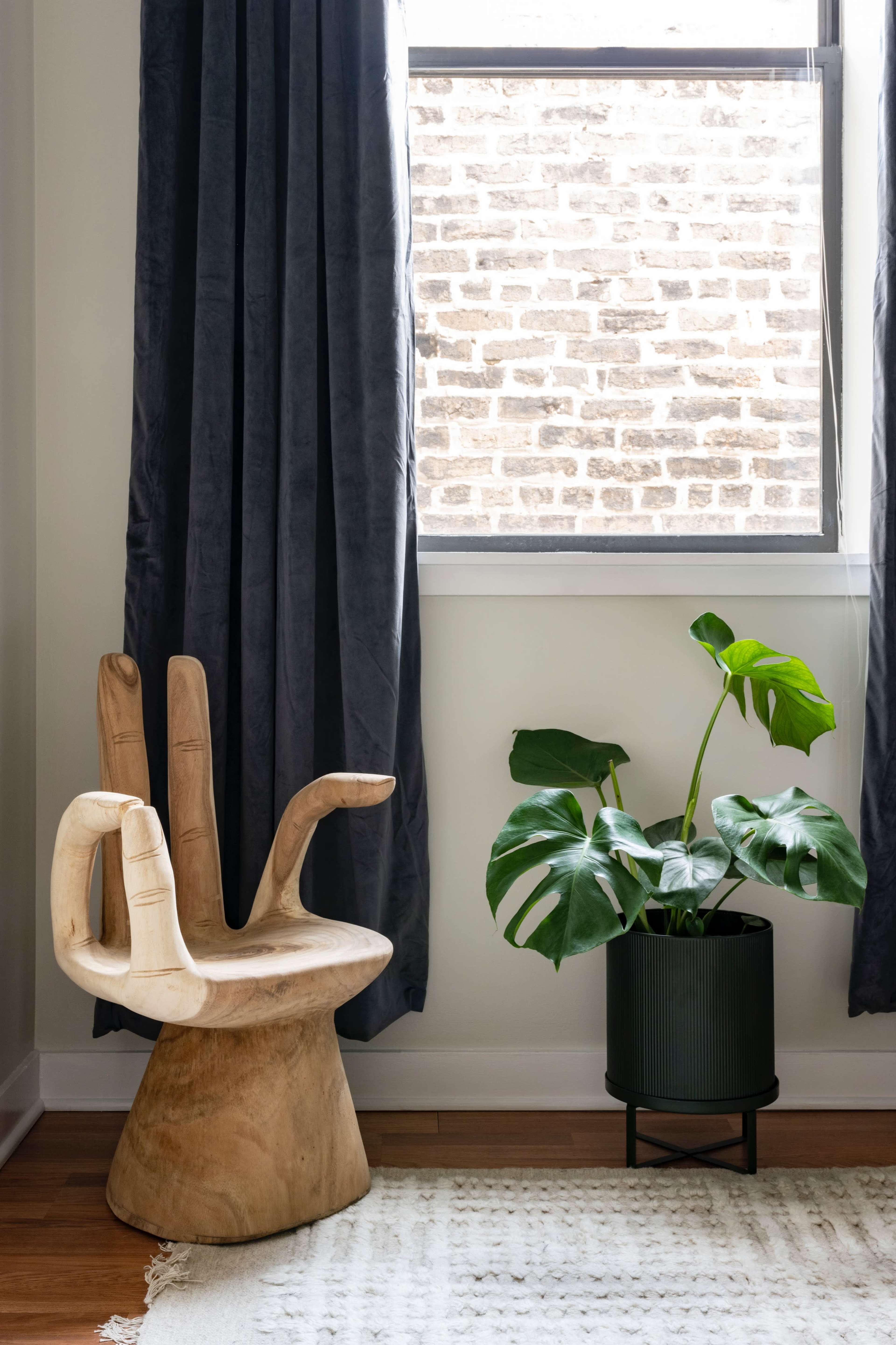 A sculptural wooden chair with hands extends from its backrest sits beside a potted monstera plant near a window covered with dark curtains.