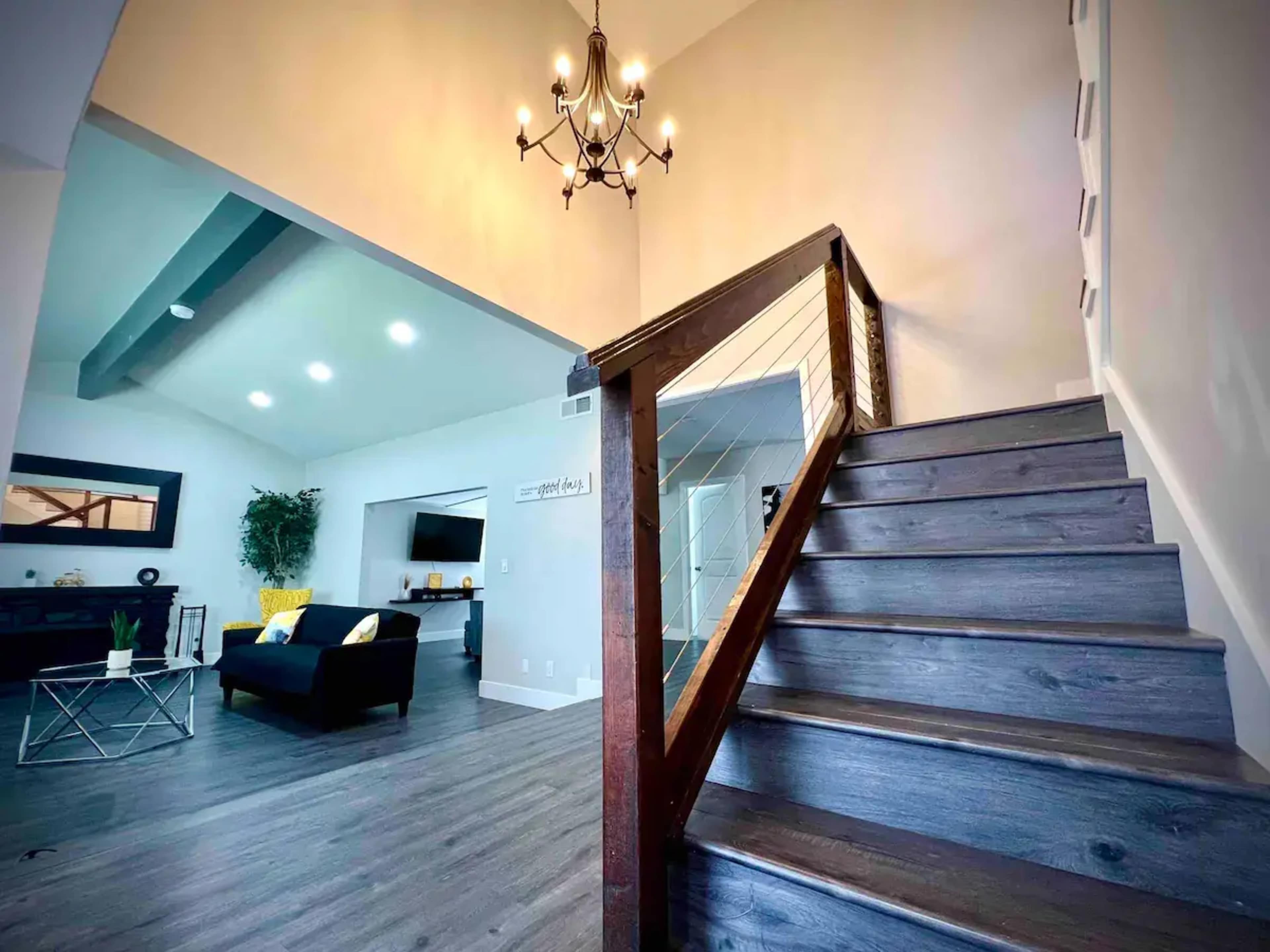 A staircase leads to a bright upper area with a modern chandelier, overlooking a spacious living room furnished with a black sofa and a television.