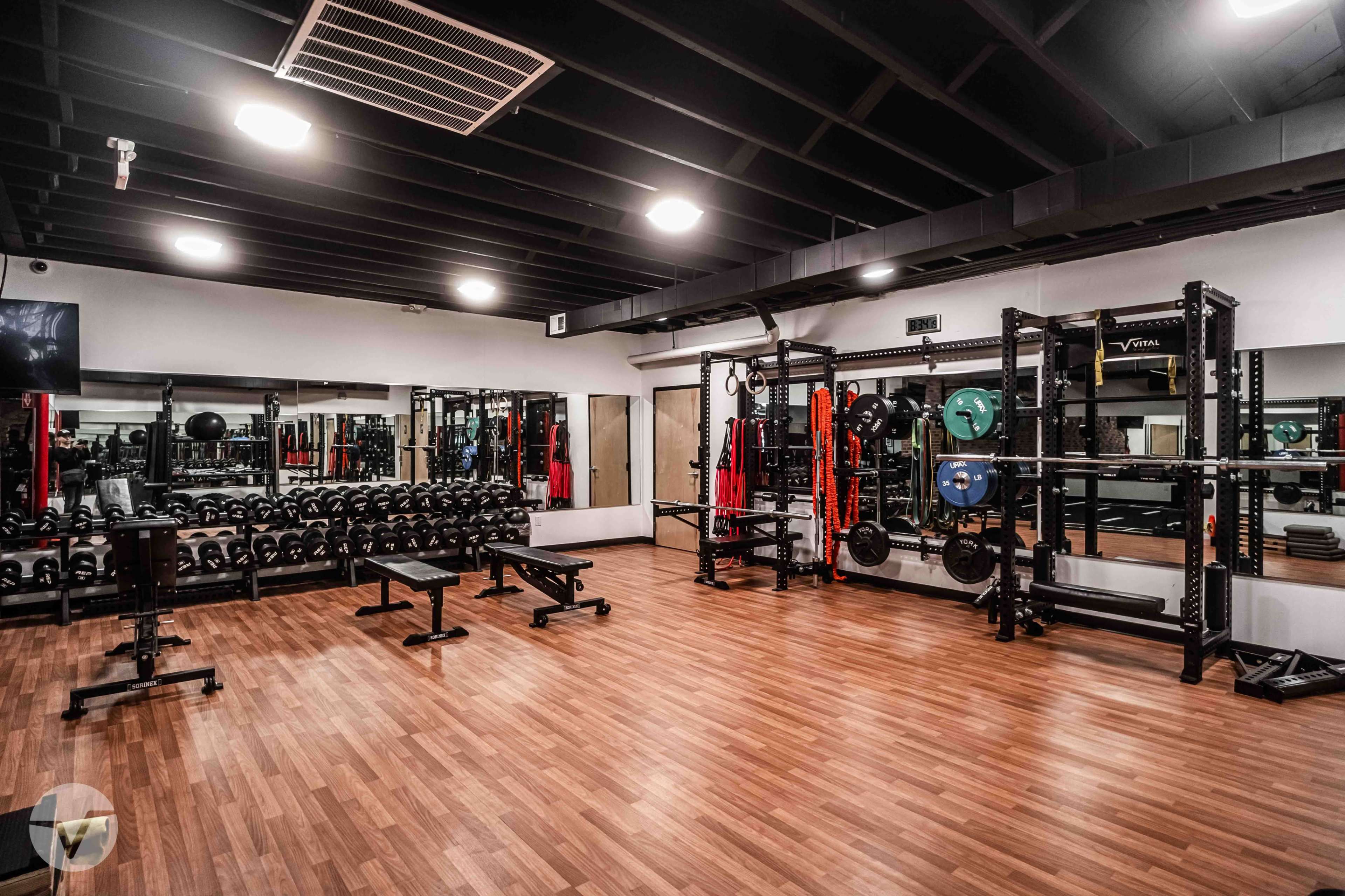 The image shows a well-equipped gym interior featuring weightlifting racks, dumbbells, and exercise mats on a wooden floor under black ceiling beams.