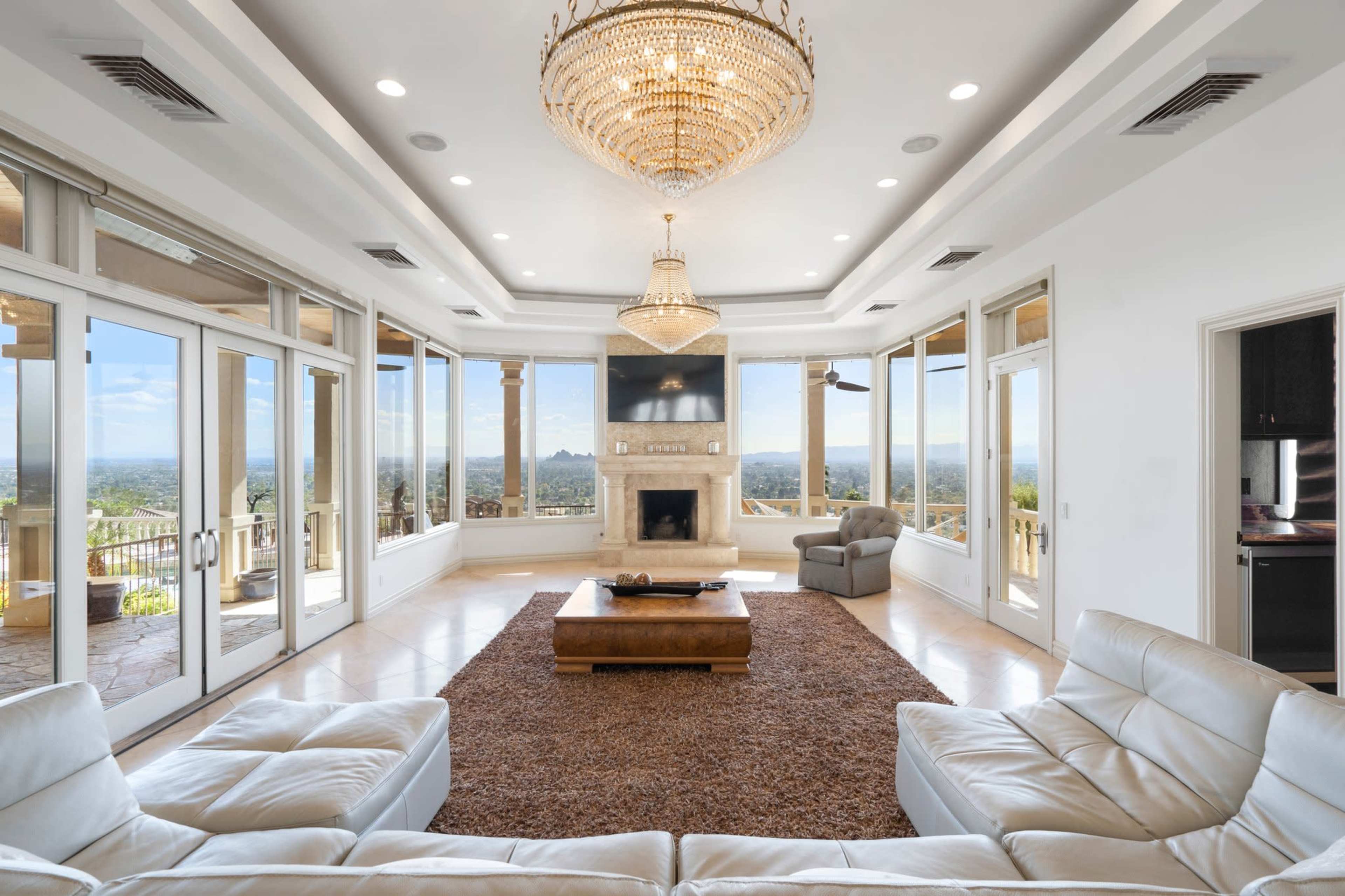 The spacious living room features large windows showcasing a panoramic view, a central fireplace, and a chandelier.