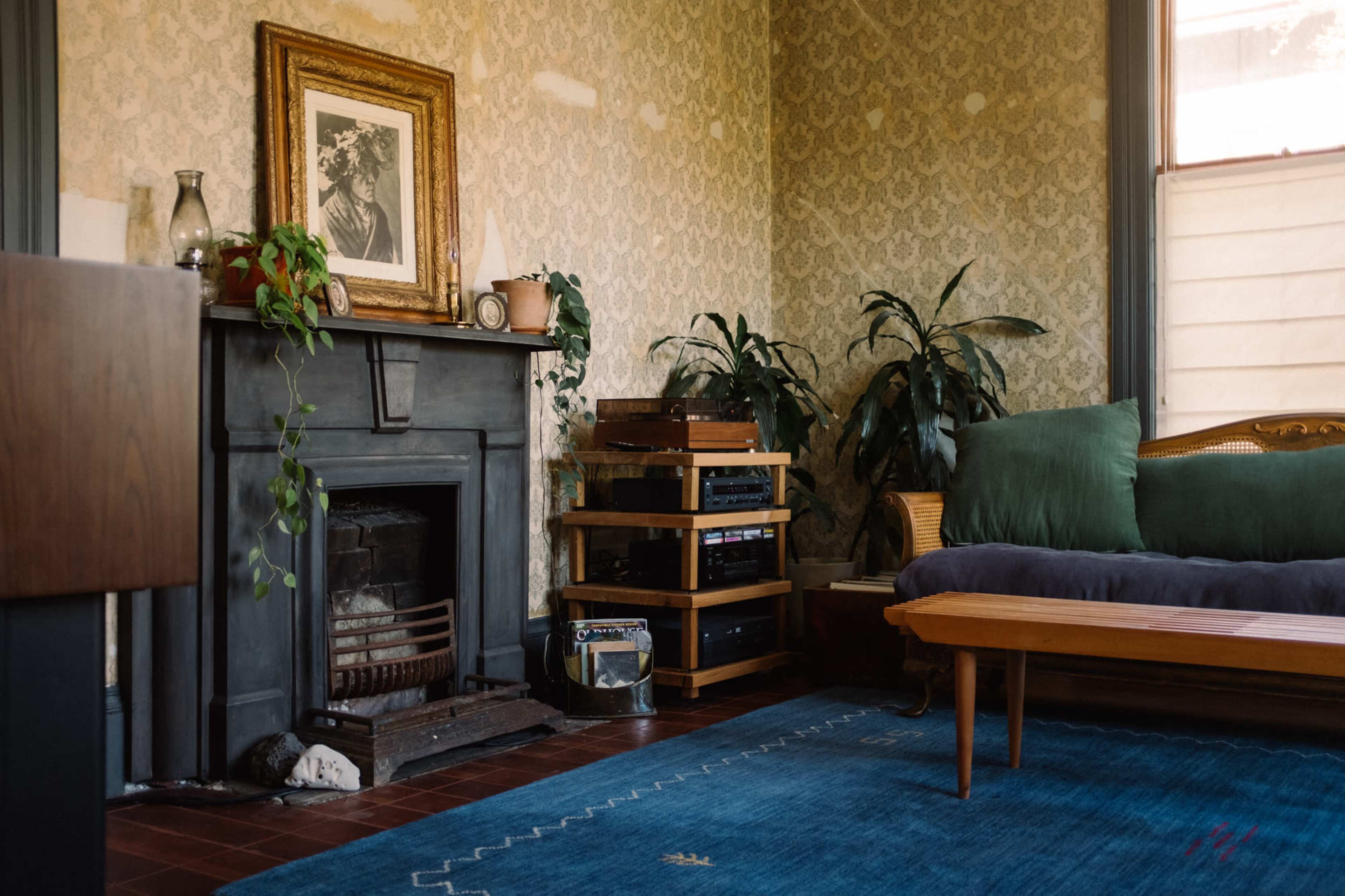 A cozy living room features a vintage fireplace, a wooden bench, and a collection of plants alongside a blue area rug.