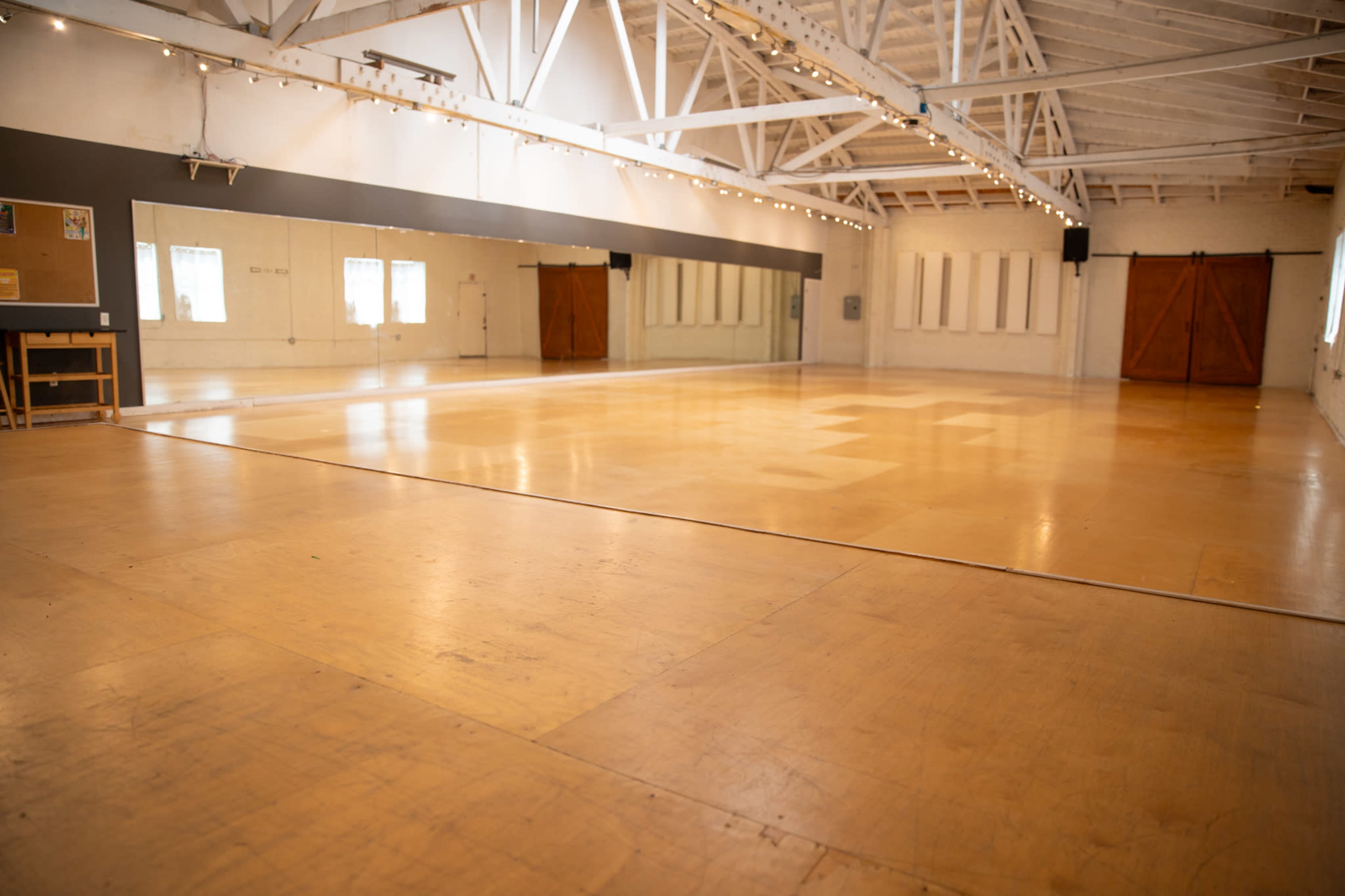 The image shows a spacious, empty wooden dance studio with large mirrors, wooden floors, and natural light streaming in through windows.
