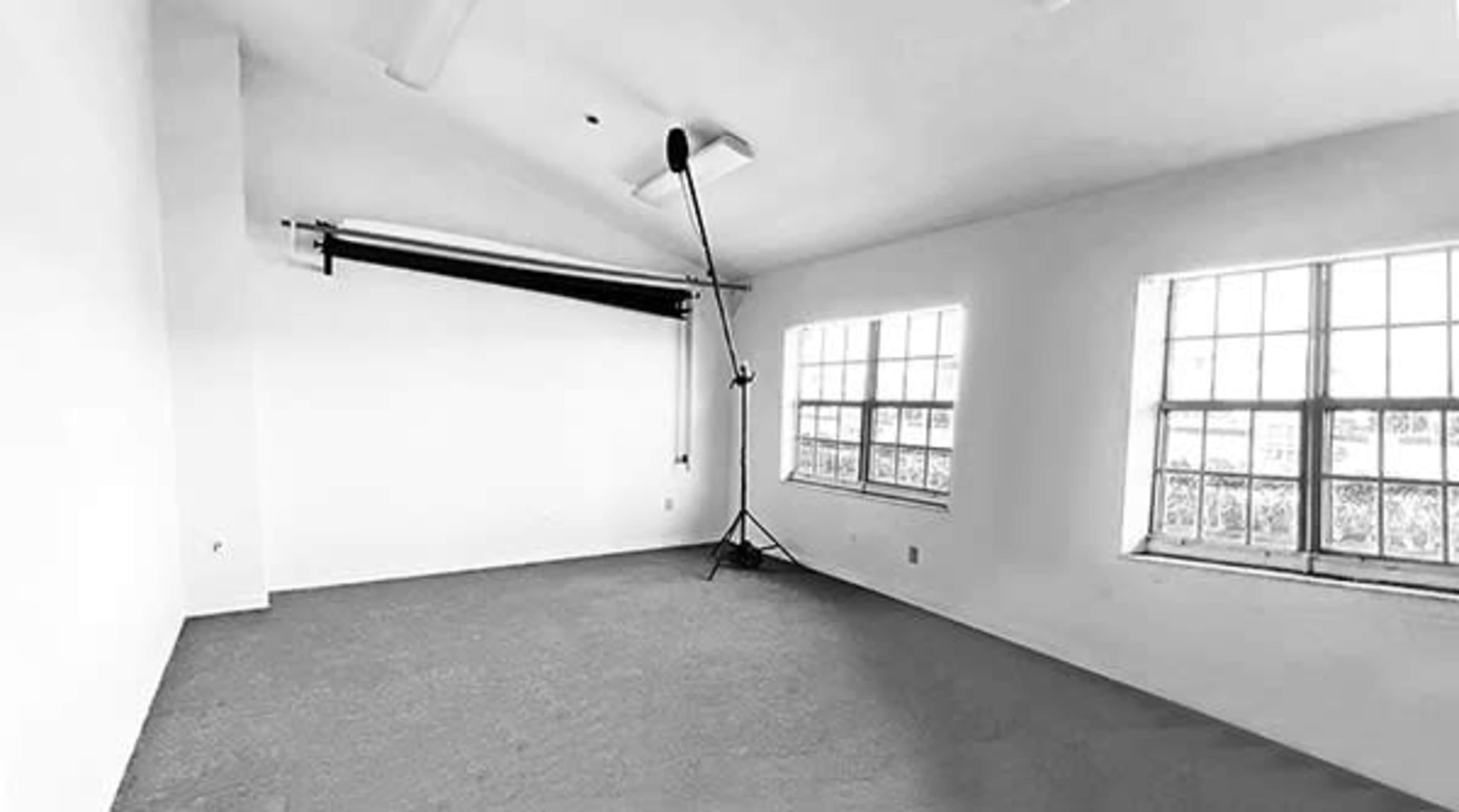 The image shows a minimalist empty room with two windows, a carpeted floor, and a backdrop stand in one corner.