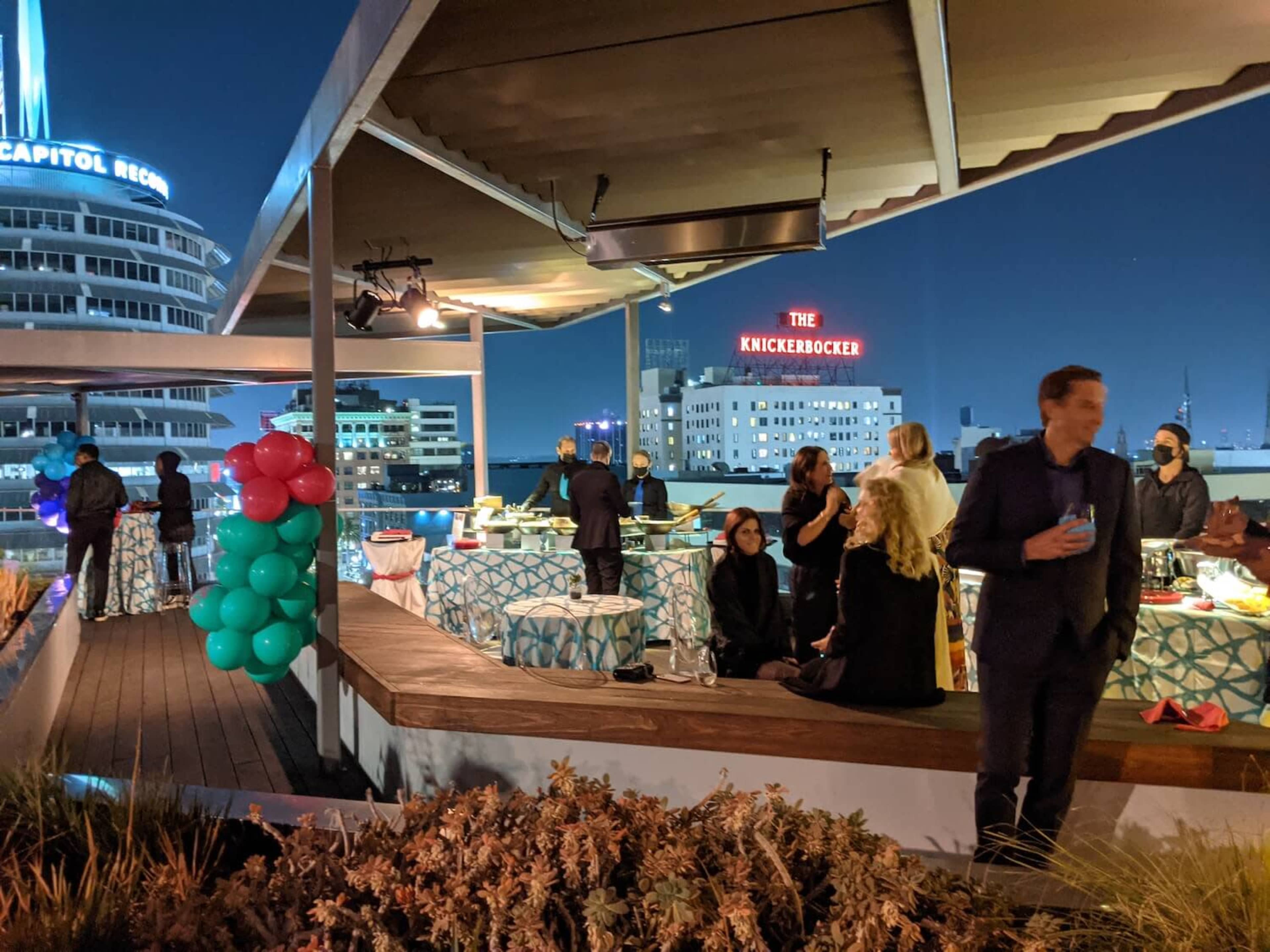 An outdoor rooftop event features guests mingling near tables decorated in turquoise and white, with city lights and a prominent sign for "The Knickerbocker" in the background.