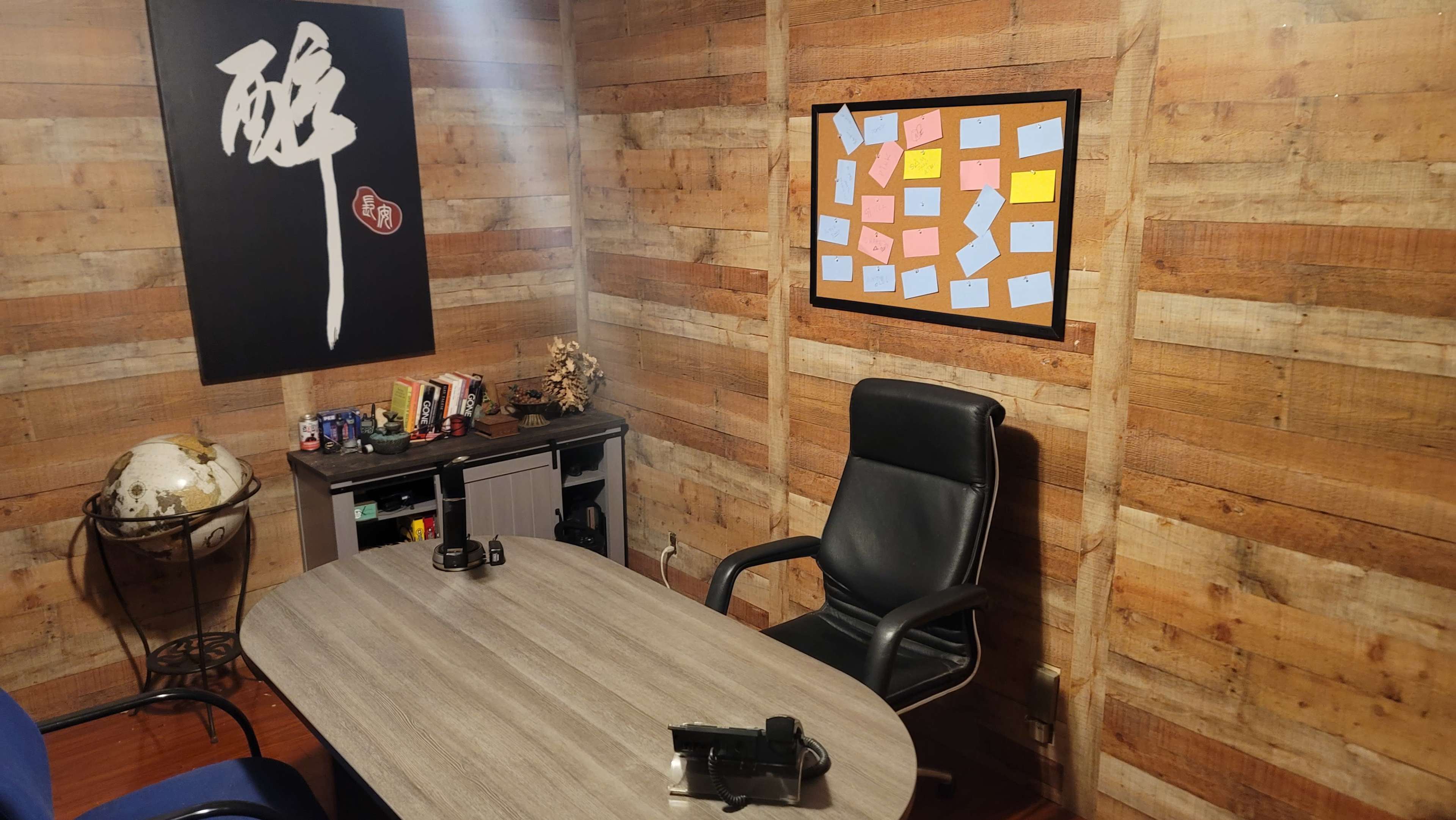 A cozy office with wooden walls, a round table, two chairs, a blackboard with colorful sticky notes, and a small shelf containing various items.