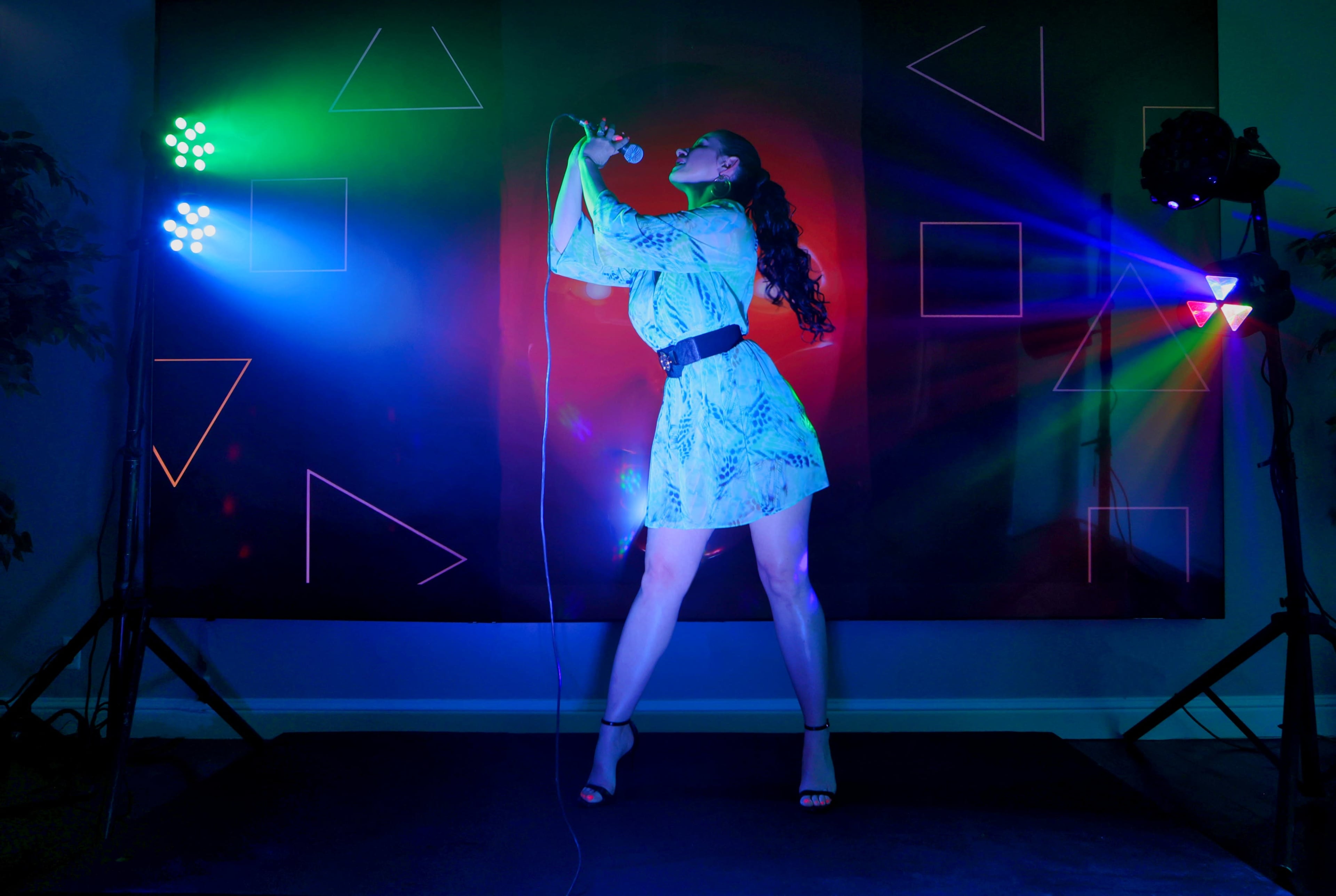 A performer stands on stage, singing into a microphone while vibrant multicolored lights and geometric shapes illuminate the background.