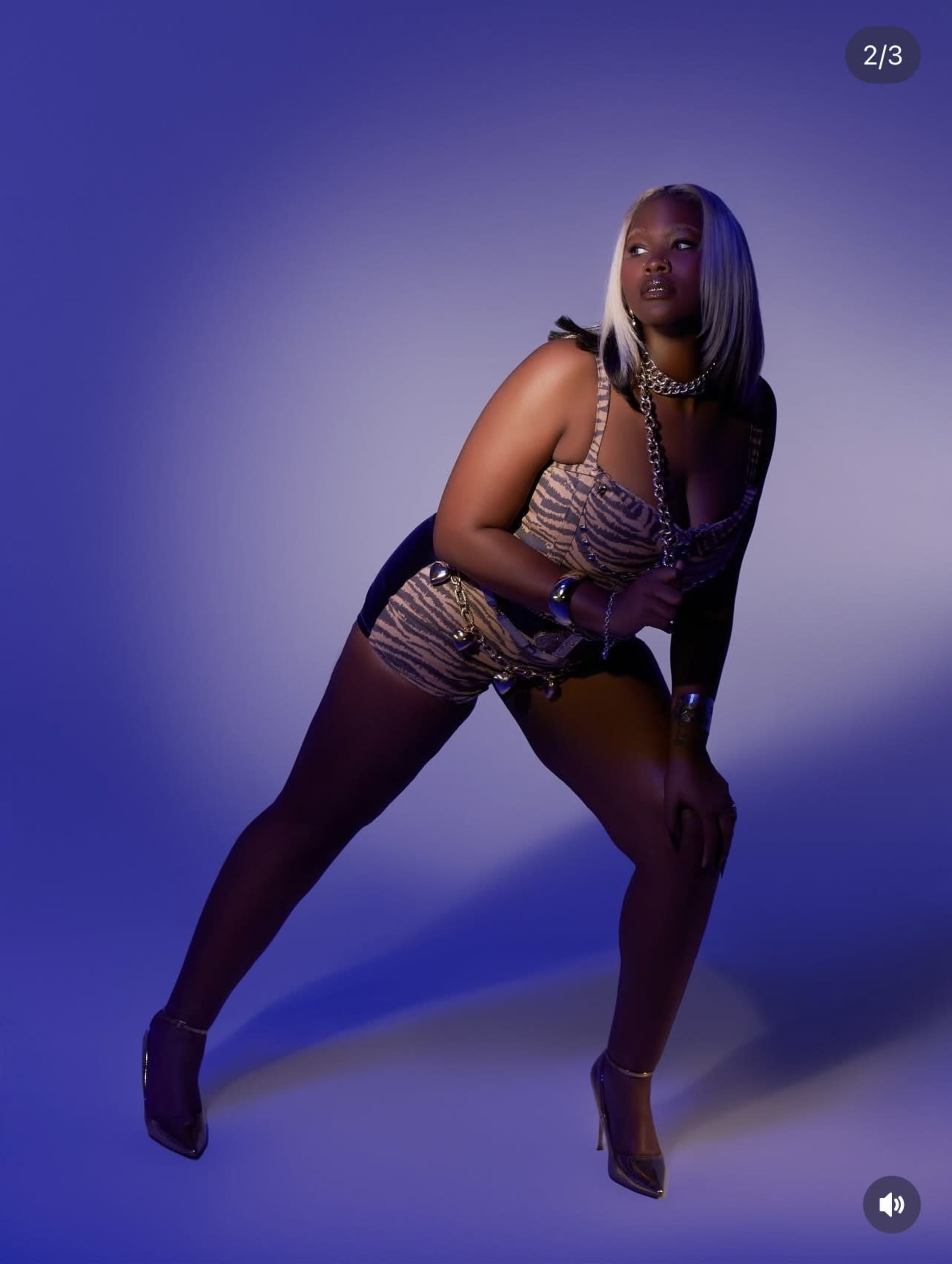 A model poses in a stylish blue and gold patterned outfit with high heels against a blue-lit background.