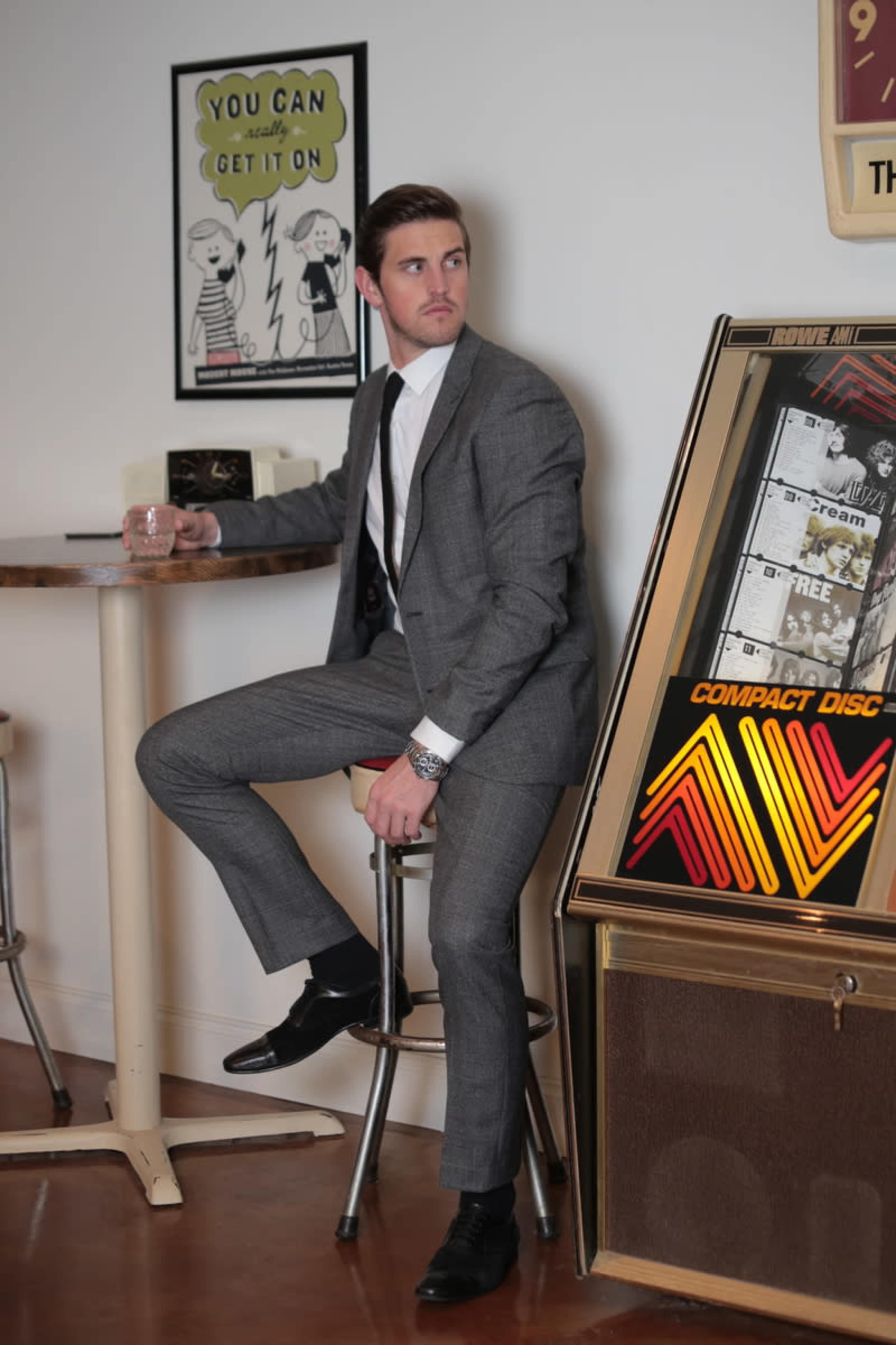 A man in a gray suit sits on a stool next to a vintage jukebox, holding a glass in one hand and looking off to the side.