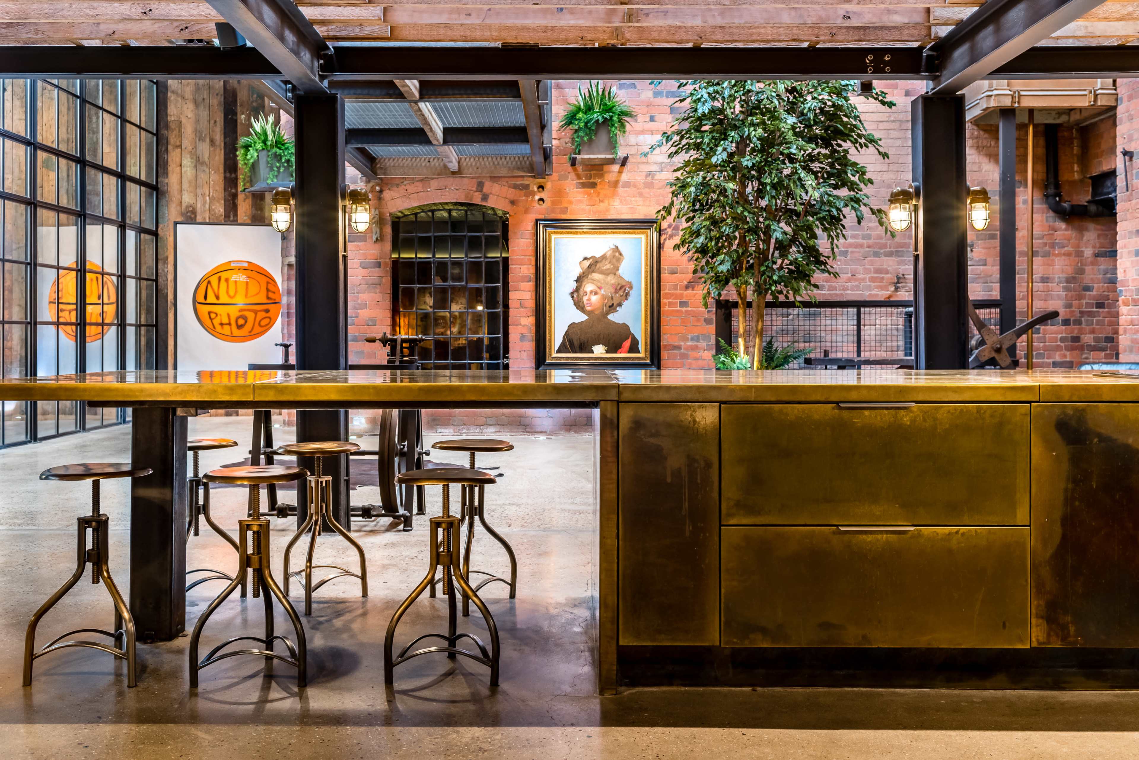 A modern industrial kitchen area with a large metallic island, wooden stools, and a vibrant artwork on the wall.