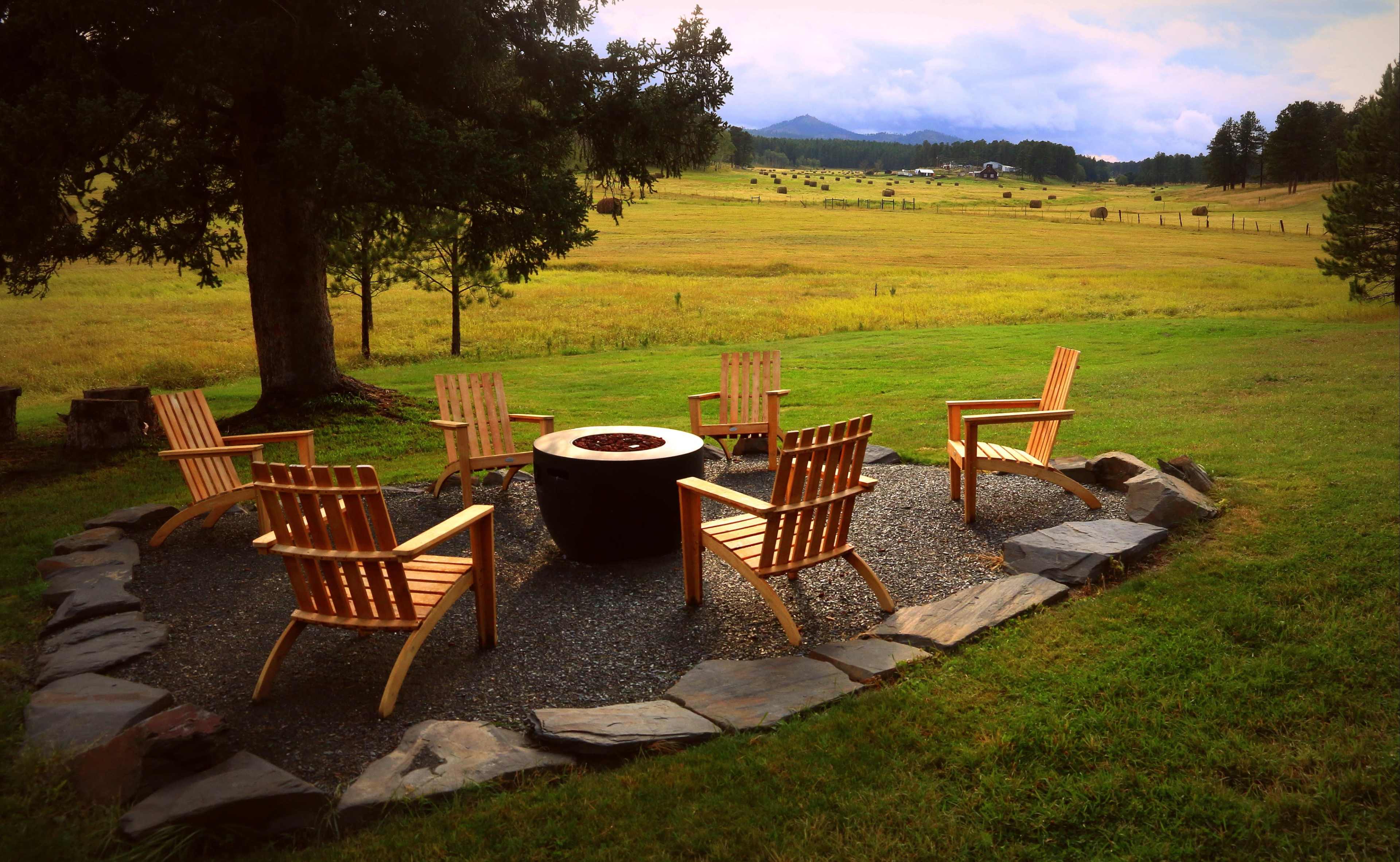A circular arrangement of wooden chairs surrounds a stone fire pit in a grassy area with a backdrop of rolling hills.