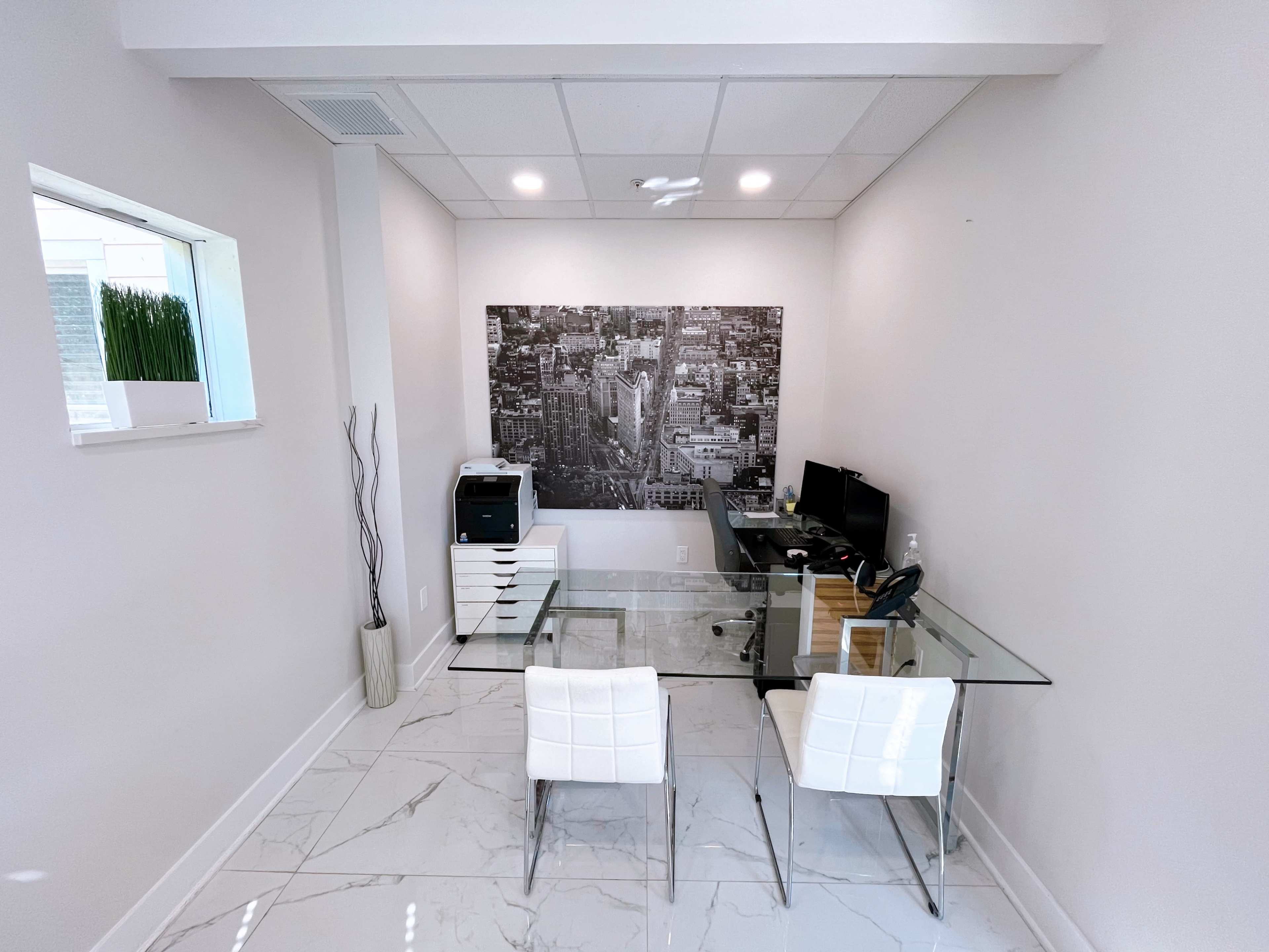 A minimalist office space with a glass desk, two white chairs, a computer setup, and a large black-and-white cityscape mural on the wall.